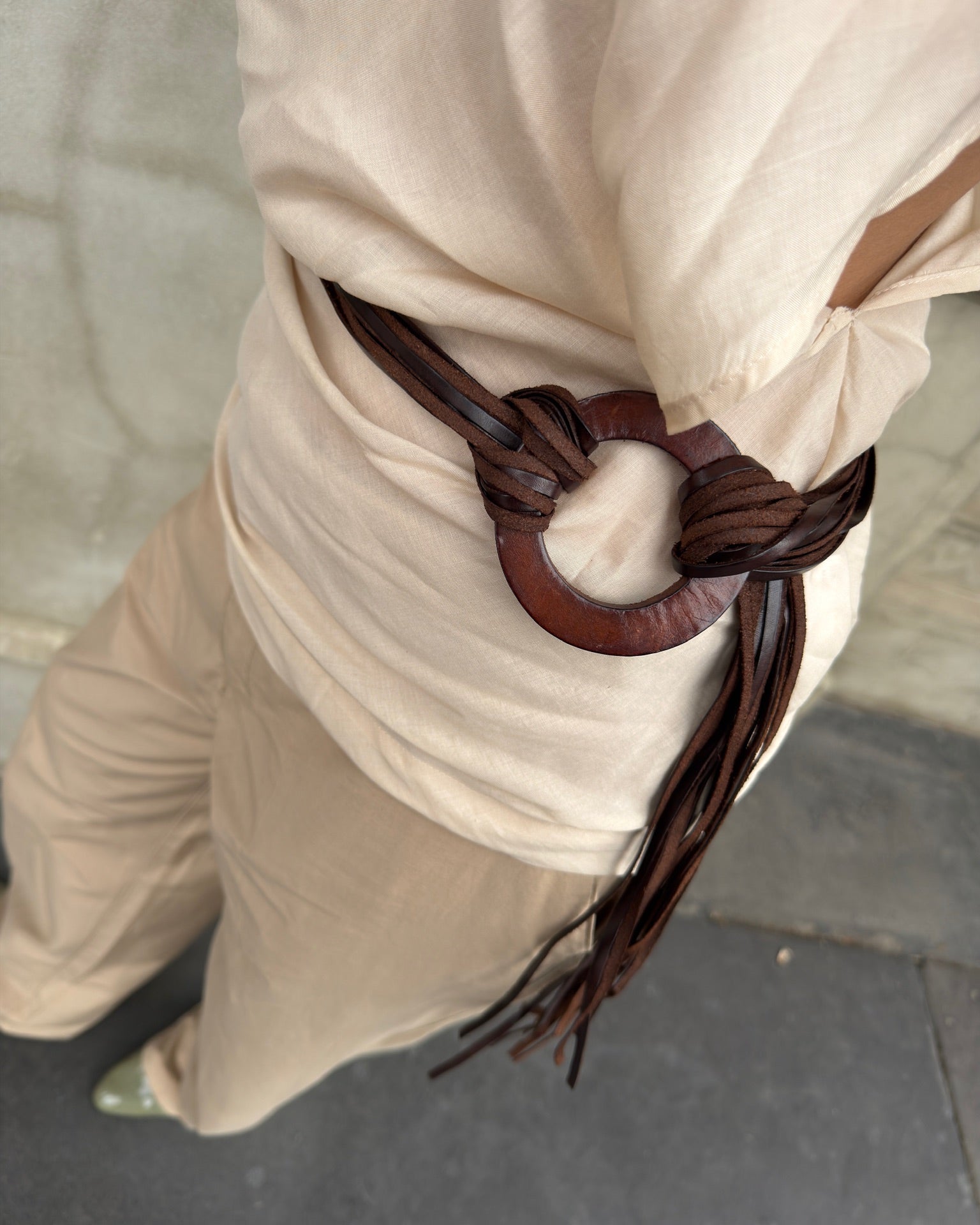 Close-up of a brown leather wrap belt with a circular wooden buckle, styled over a draped beige sleeveless top and beige pants.