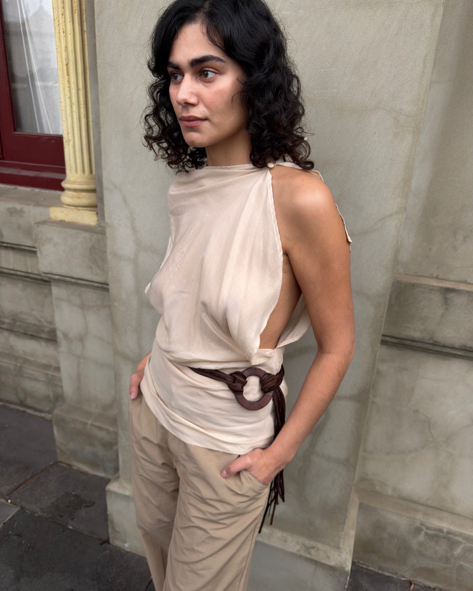 Model wearing a draped beige sleeveless top with an asymmetric open side, cinched with a brown leather belt and paired with beige trousers.