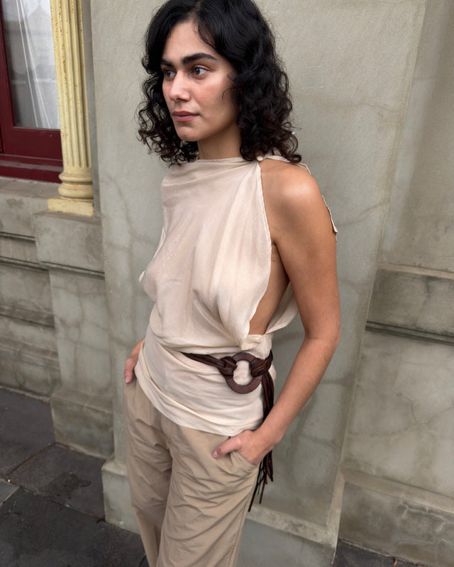Model wearing a draped beige sleeveless top with an asymmetric open side, cinched with a brown leather belt and paired with beige trousers.