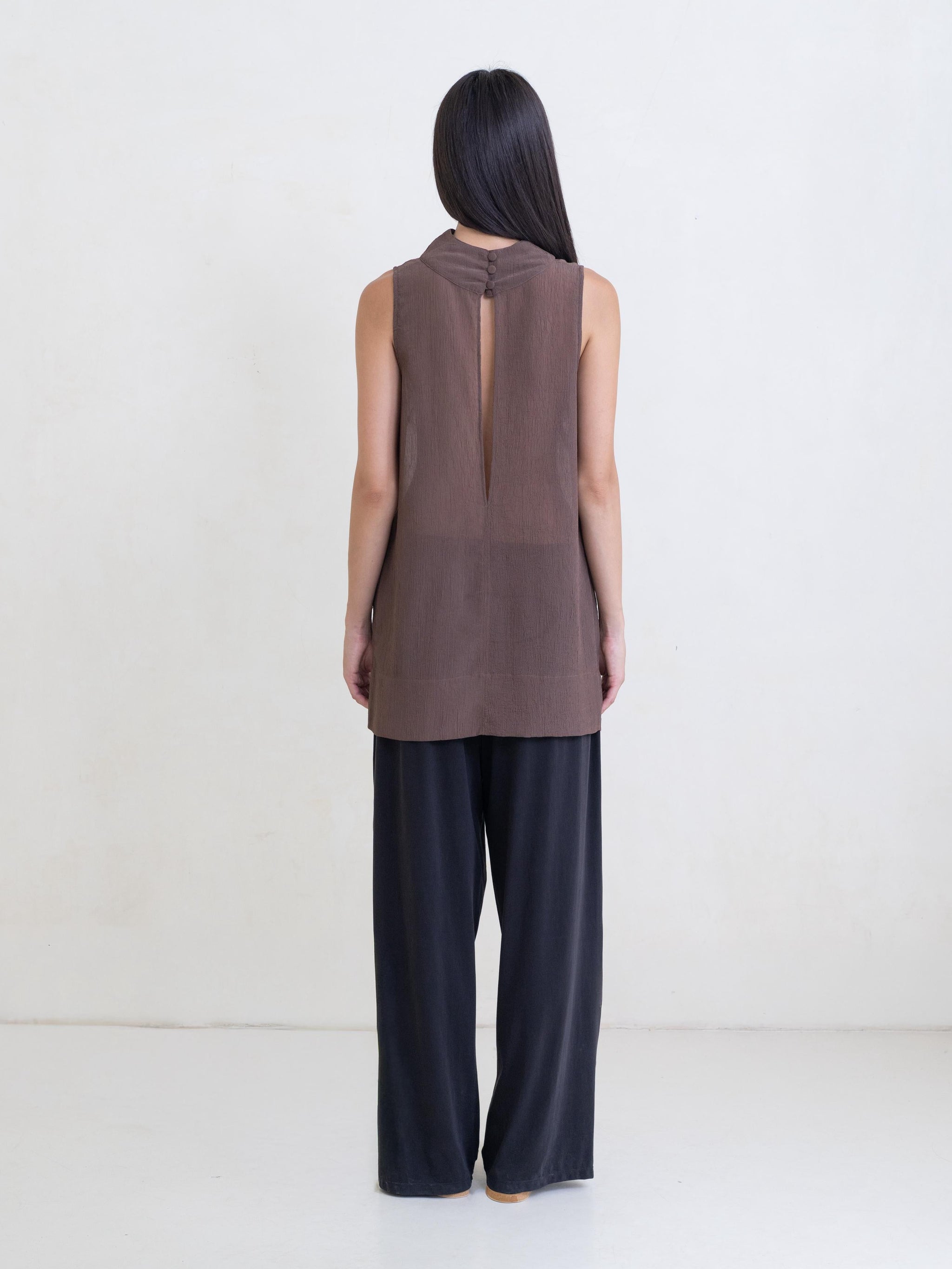 Woman wearing a sheer brown sleeveless tunic and black pants on a white background.
