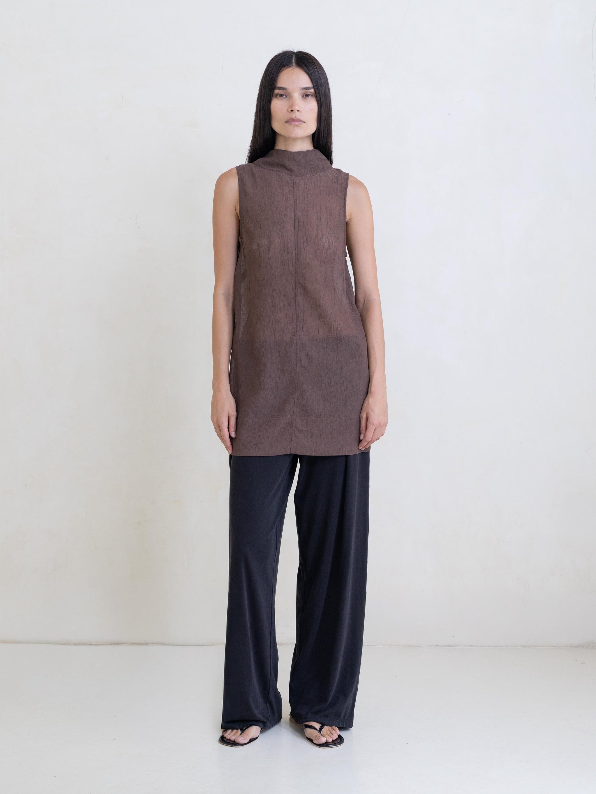 Woman wearing a sheer brown sleeveless tunic and black pants on a white background.