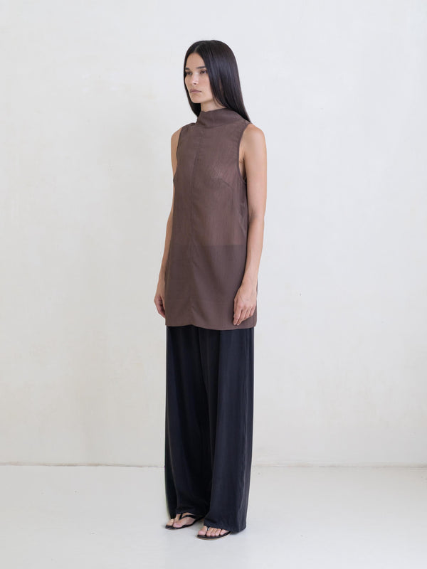 Woman wearing a sheer brown sleeveless tunic and black pants on a white background.