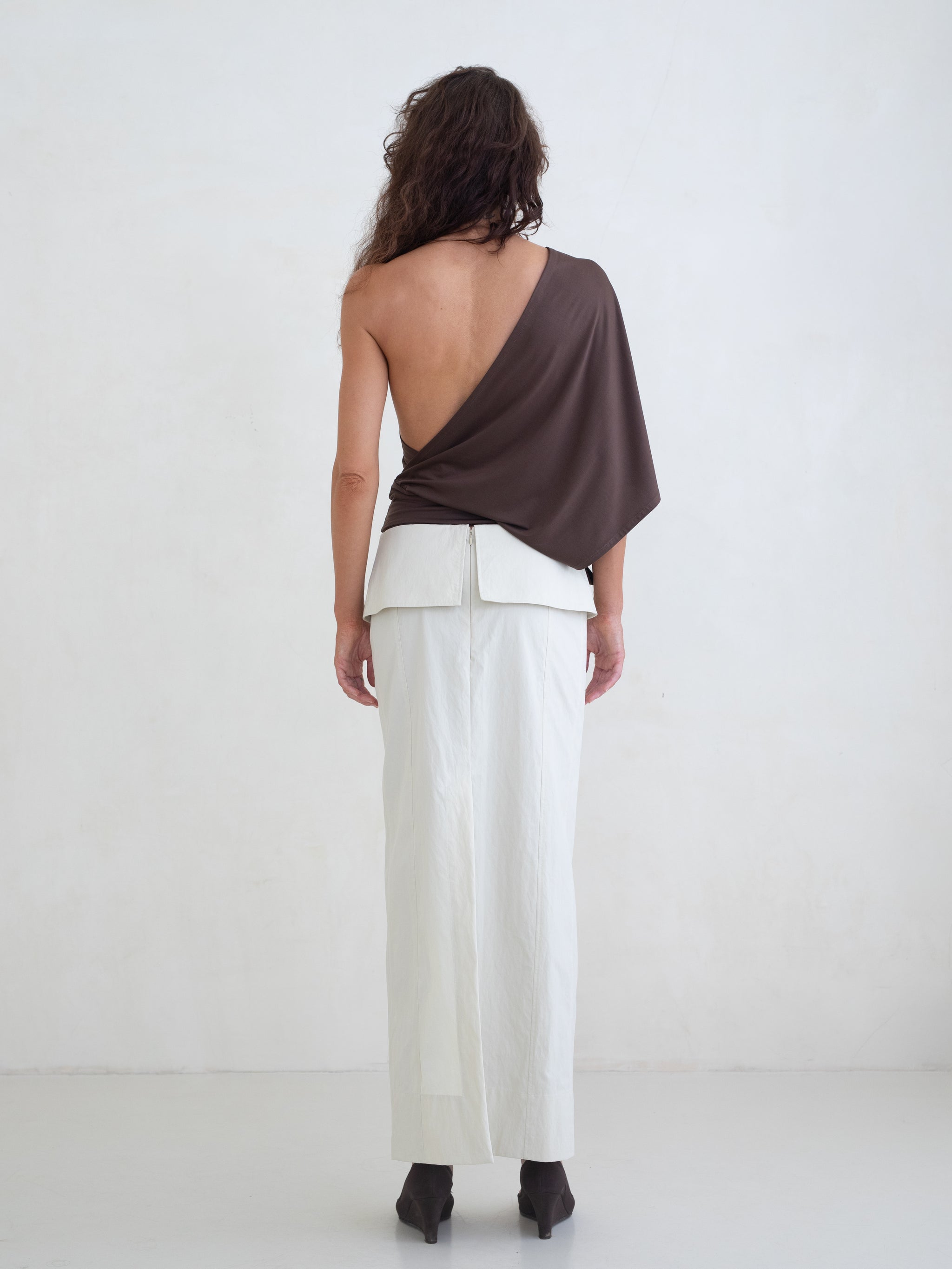 Woman wearing a brown one-shoulder top and an off-white maxi skirt on a white background. 