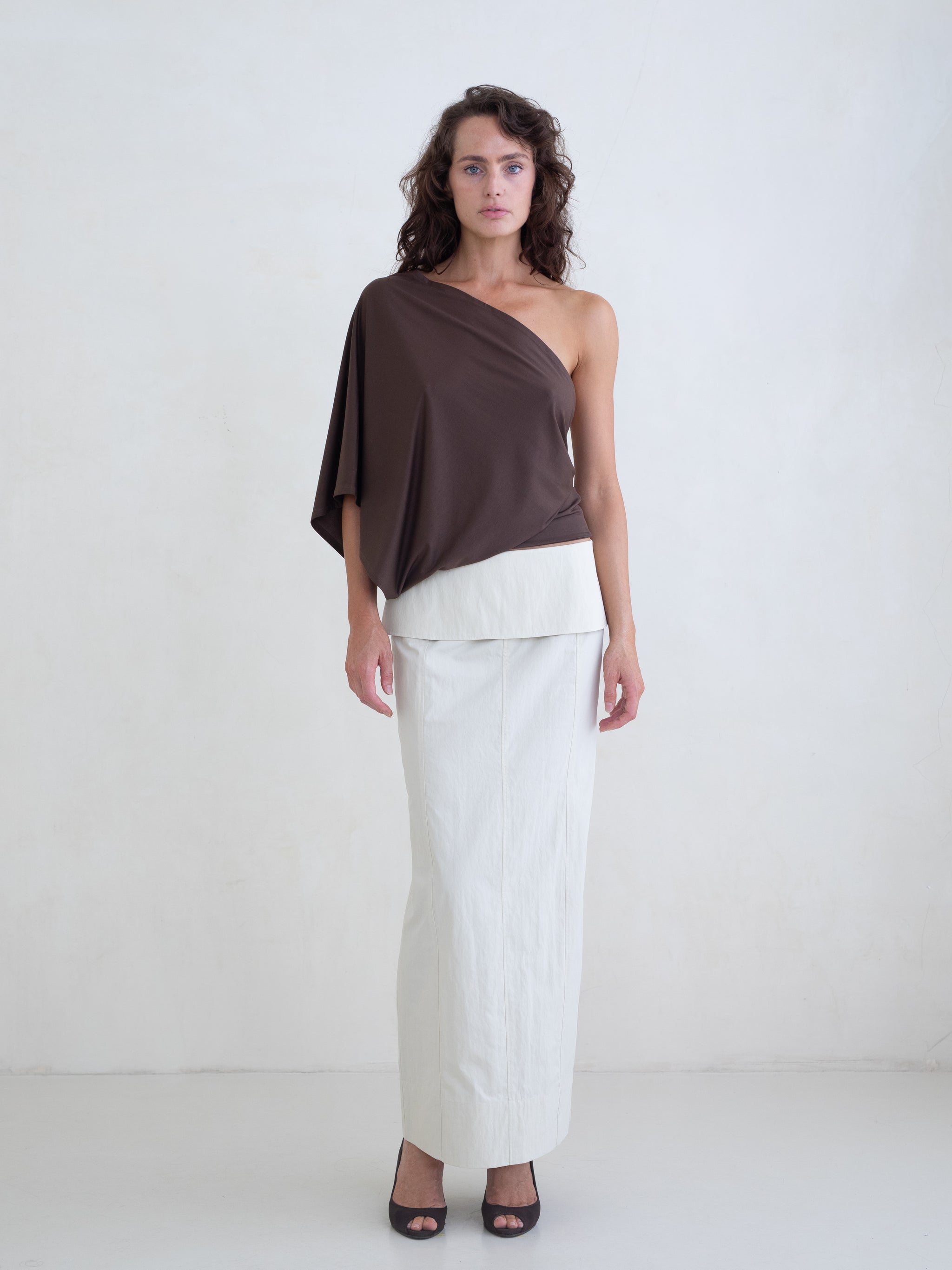 Woman wearing a brown one-shoulder top and an off-white maxi skirt on a white background. 