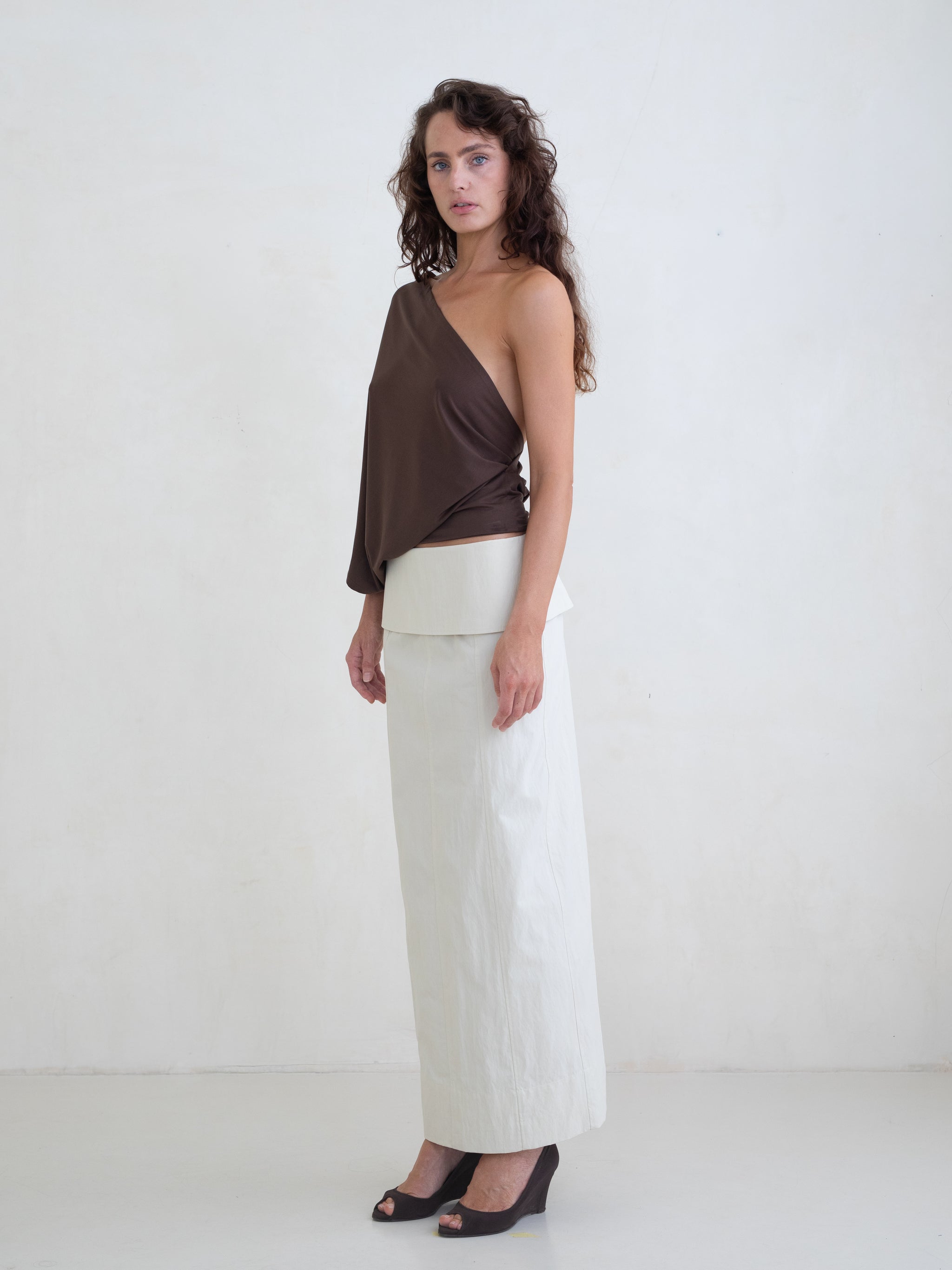 Woman wearing a brown one-shoulder top and an off-white maxi skirt on a white background. 
