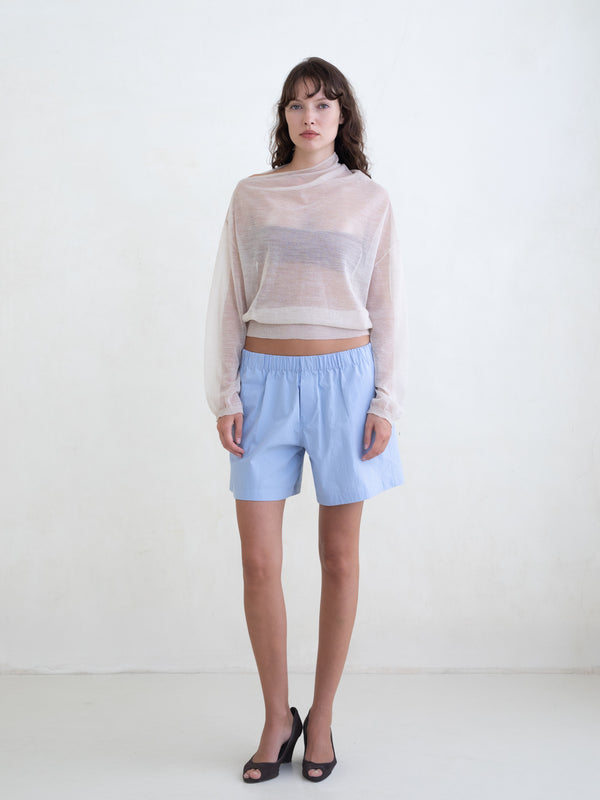 Woman wearing a sheer cream sweater and light blue boxer shorts on a white background. 