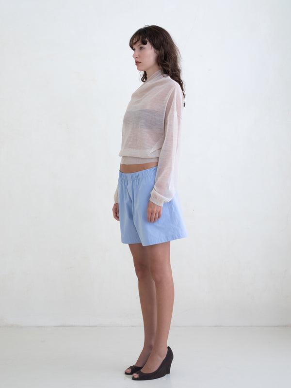 Woman wearing a sheer cream sweater and light blue boxer shorts on a white background. 