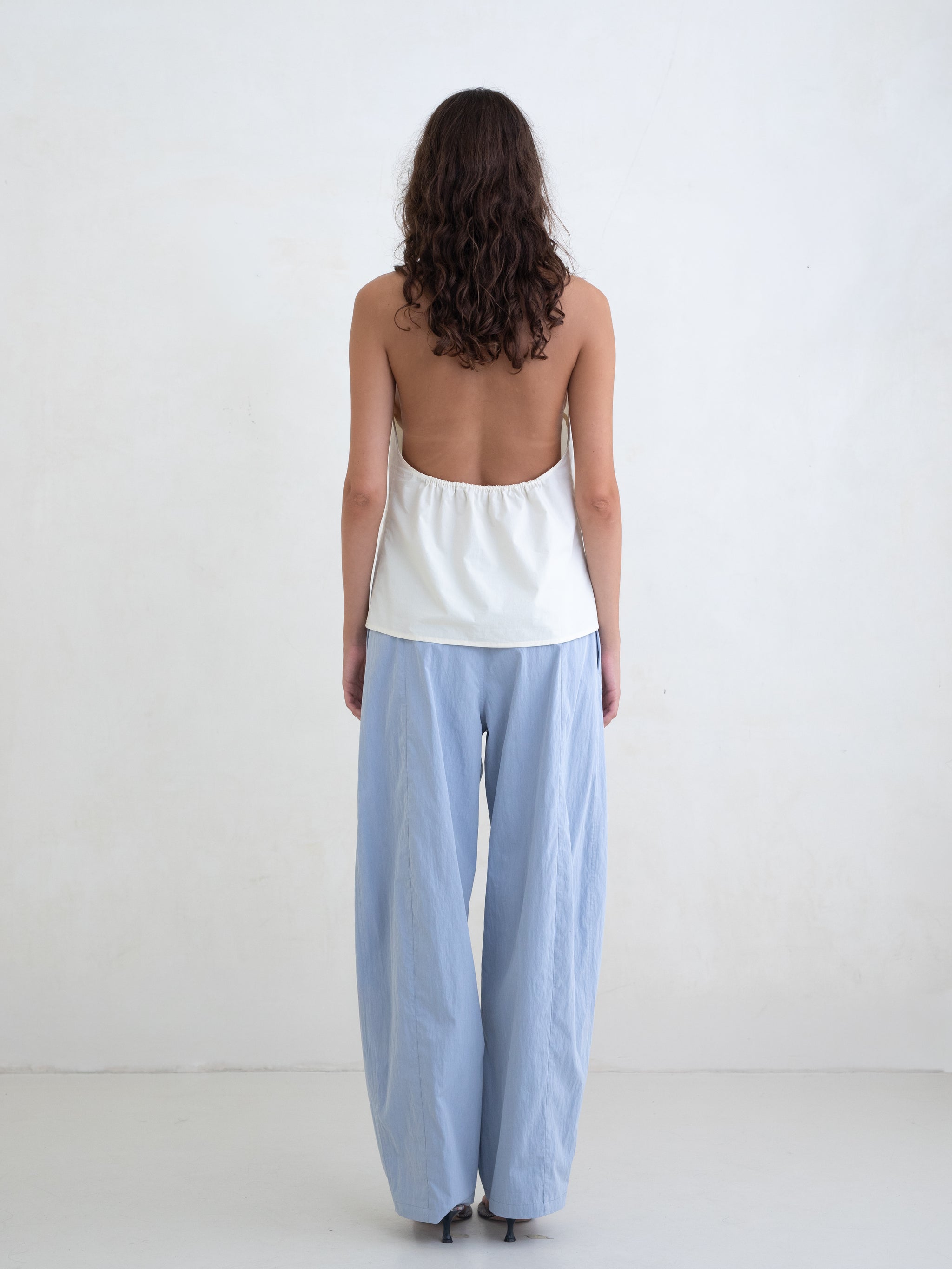 Woman wearing a cream v-neck halter top and light blue straight leg trousers on a white background. 