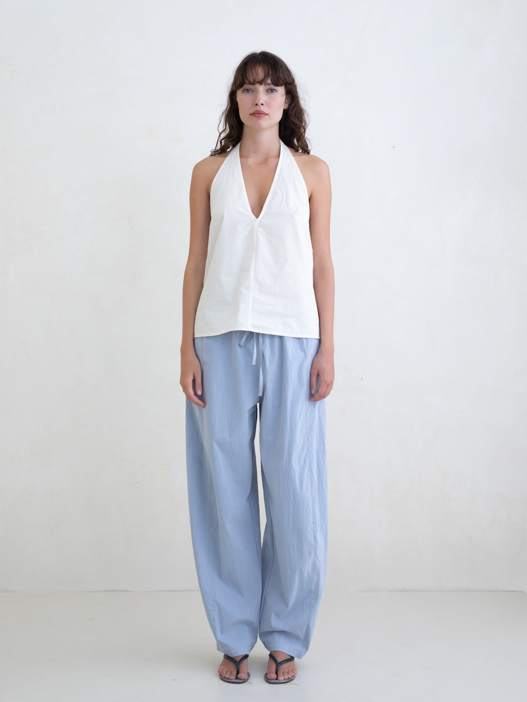 Woman wearing a cream v-neck halter top and light blue straight leg trousers on a white background. 