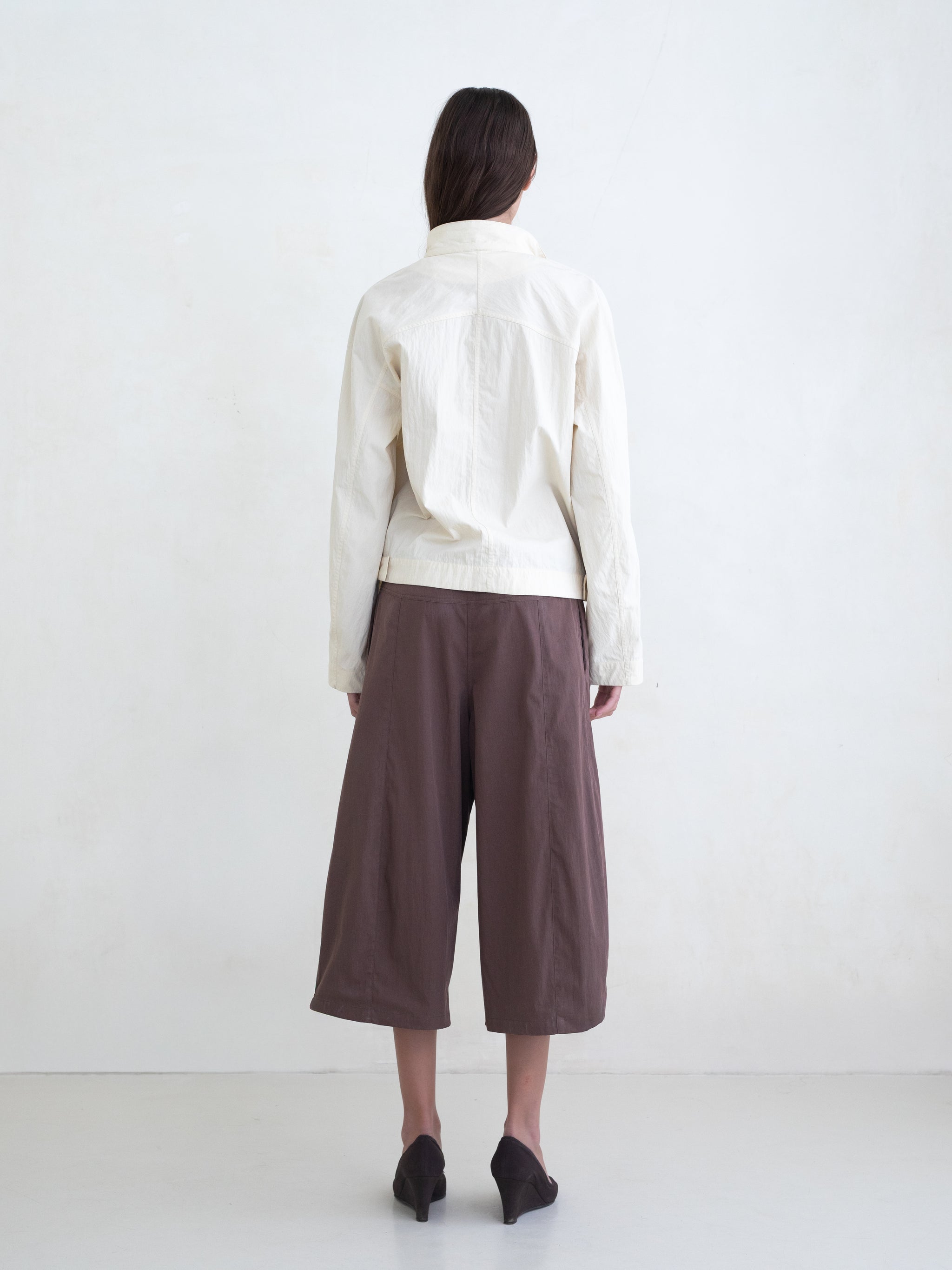 Woman wearing a cream asymmetric jacket and brown culotte pants on a white background. 