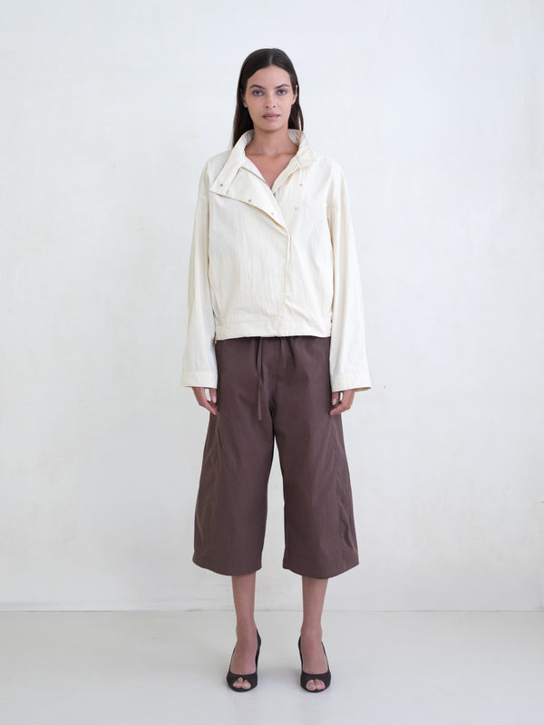Woman wearing a cream asymmetric jacket and brown culotte pants on a white background. 