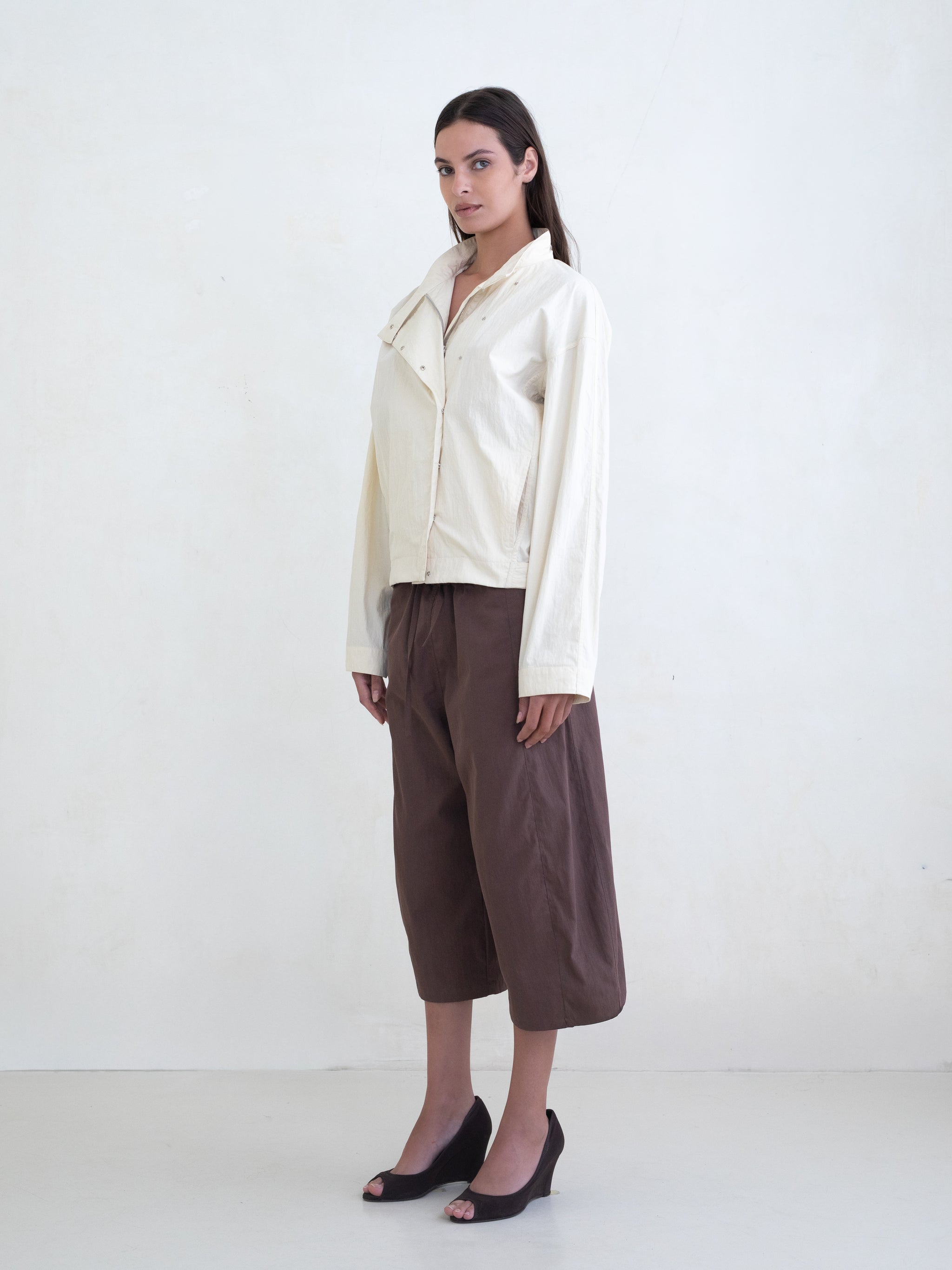 Woman wearing a cream asymmetric jacket and brown culotte pants on a white background. 