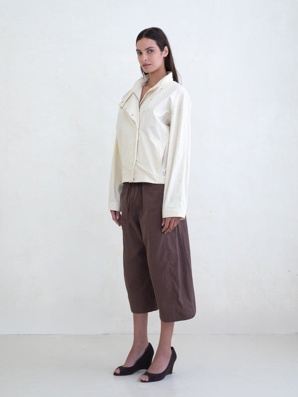 Woman wearing a cream asymmetric jacket and brown culotte pants on a white background. 