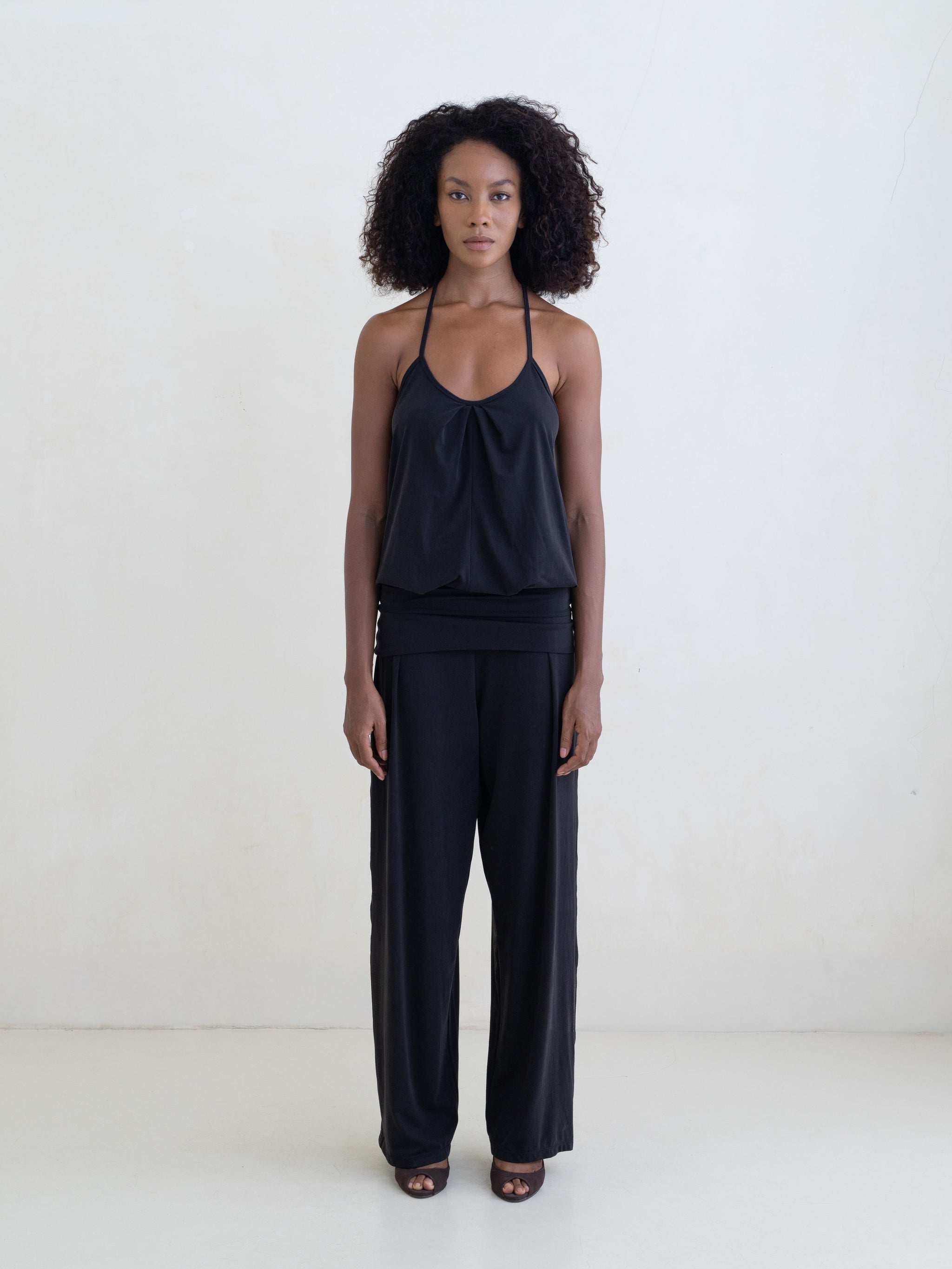 Woman wearing a black halter neck tank top and black straight leg pants on a white background. 