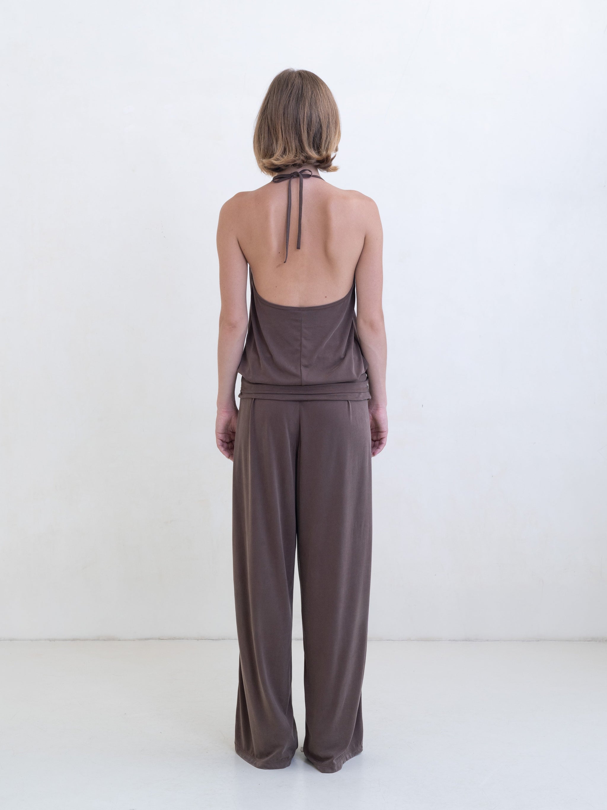 Woman wearing a brown halter neck tank top and brown straight leg pants on a white background. 