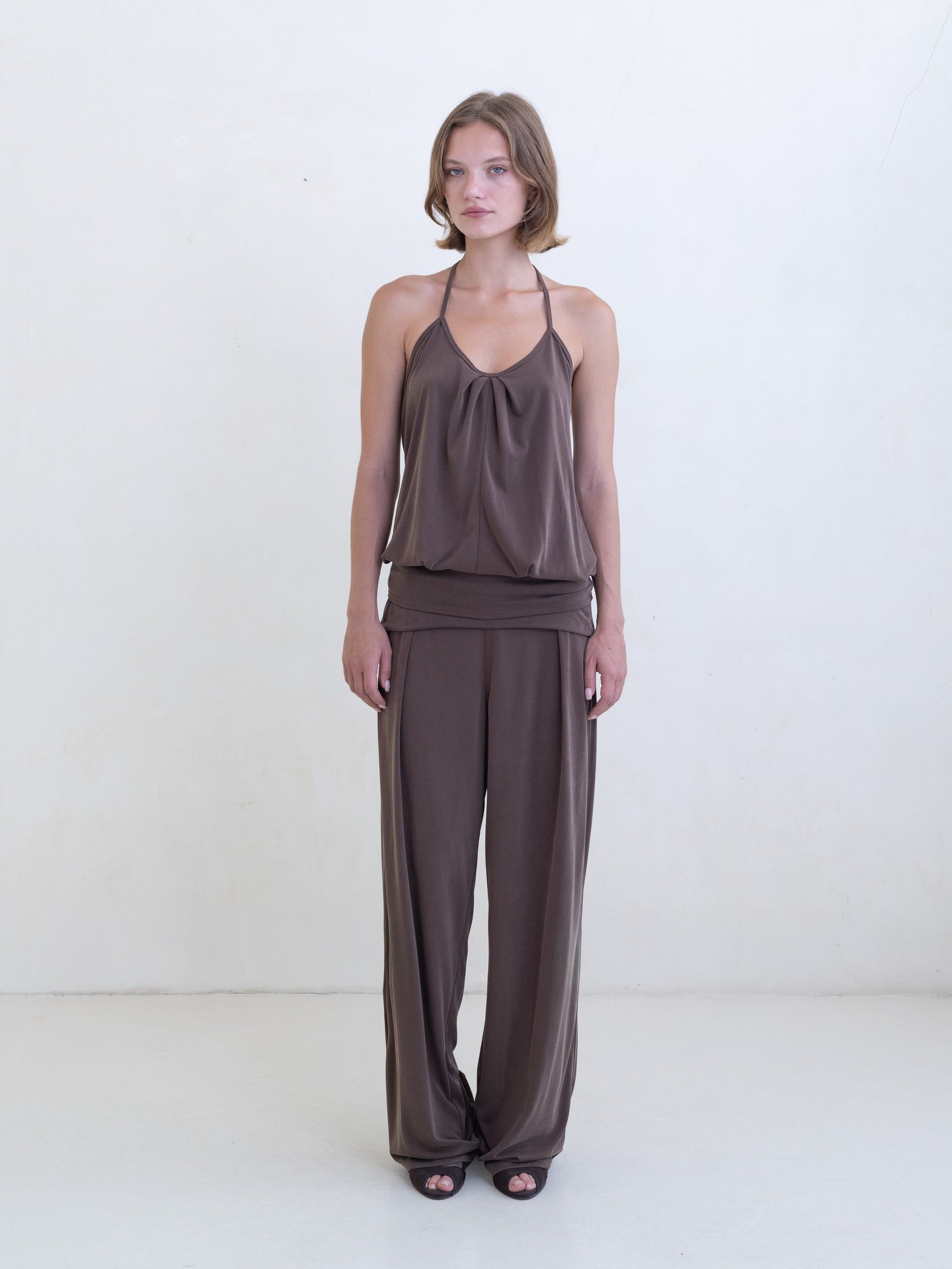 Woman wearing a brown halter neck tank top and brown straight leg pants on a white background. 