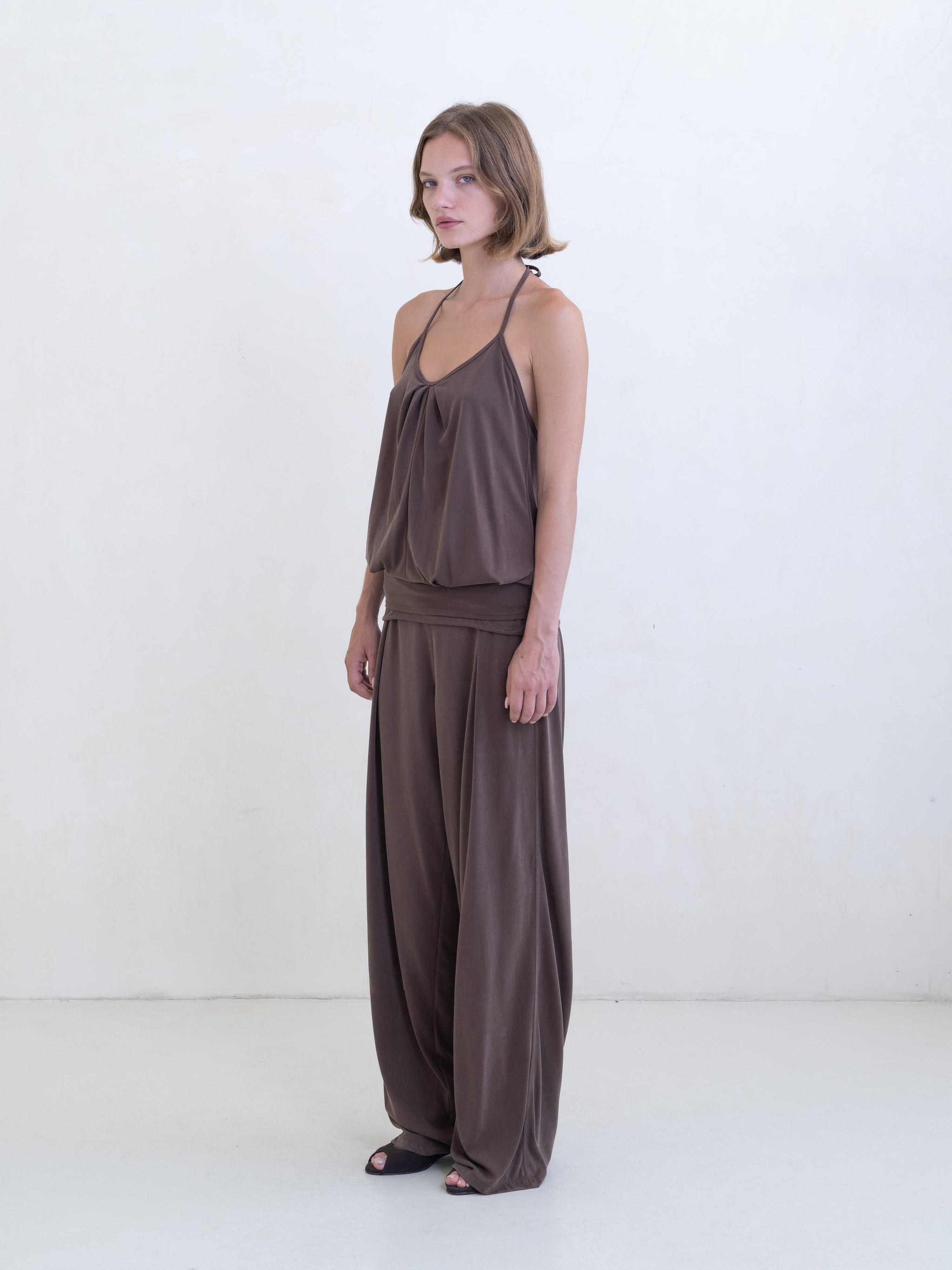 Woman wearing a brown halter neck tank top and brown straight leg pants on a white background. 