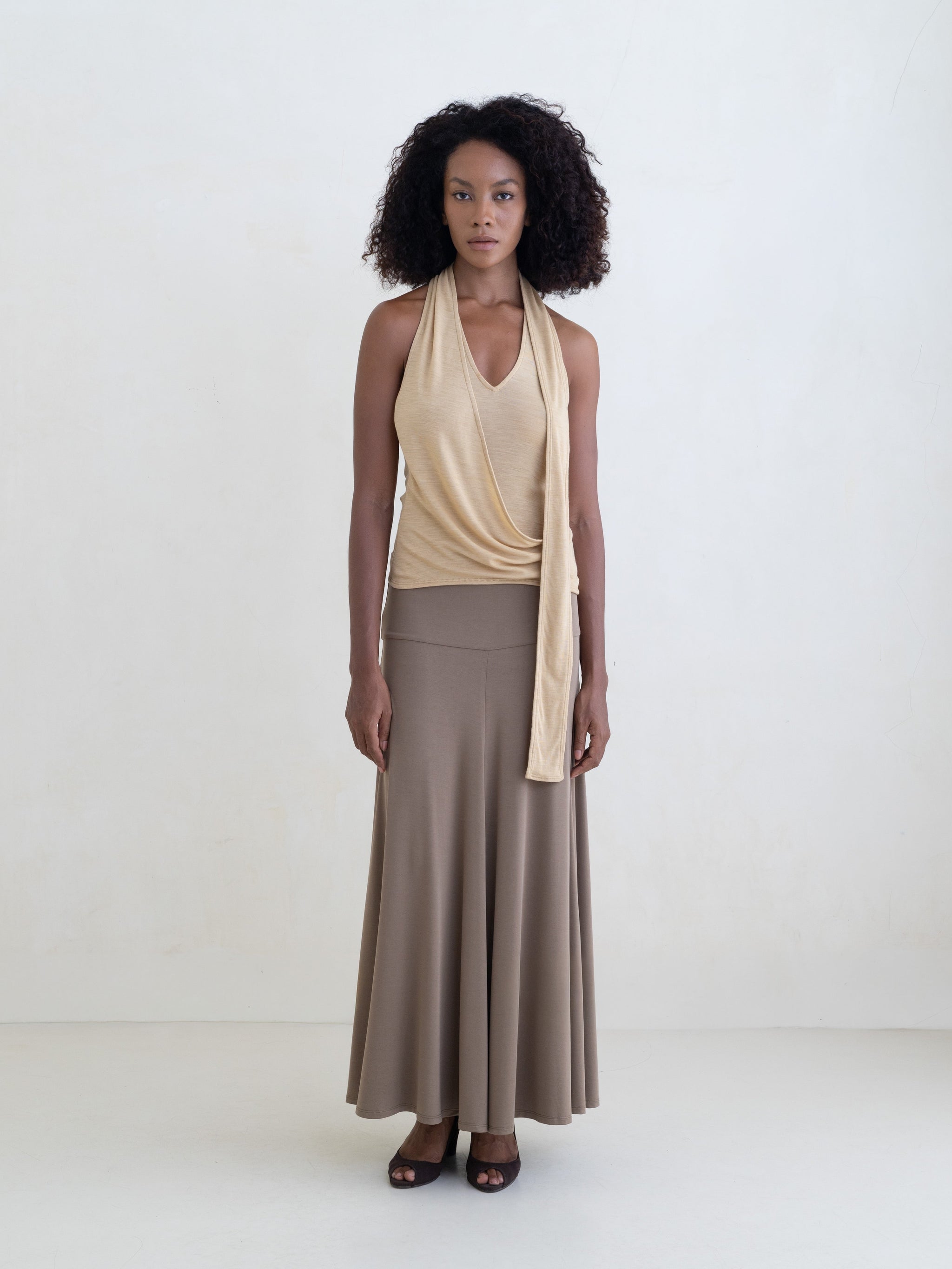 Woman wearing a light yellow sleeveless halter-style wrap top and a taupe maxi skirt on a white background. 