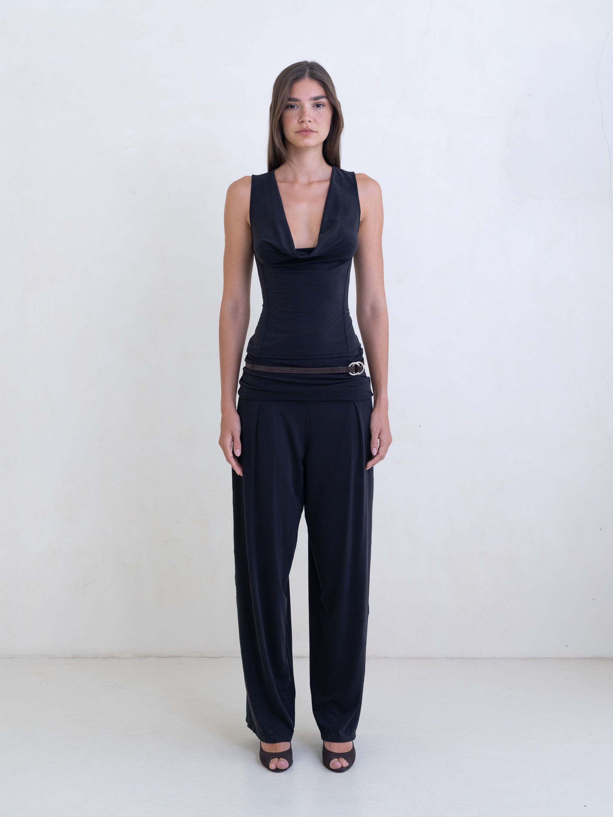 Woman wearing a black sleeveless cowl neck top and black pants on a white background. 