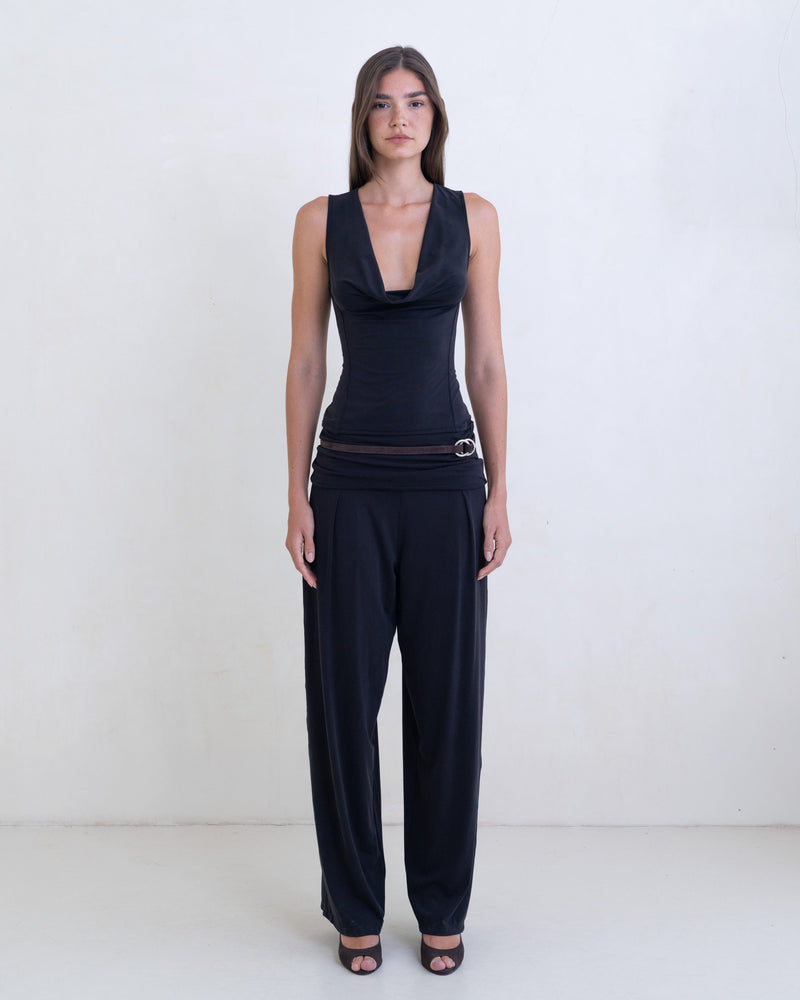 Woman wearing a black sleeveless cowl neck top and black pants on a white background. 