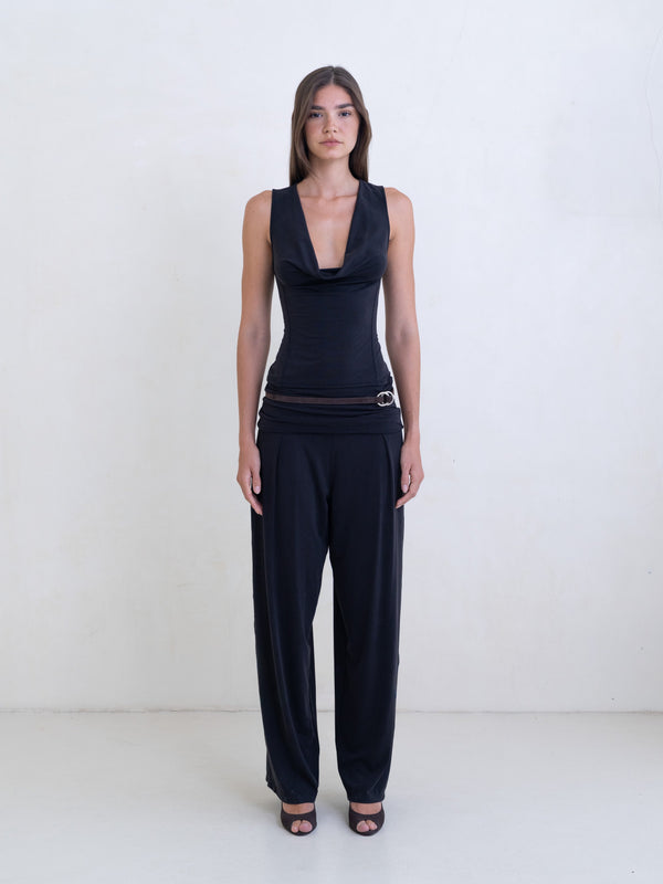 Woman wearing a black sleeveless cowl neck top and black pants on a white background. 