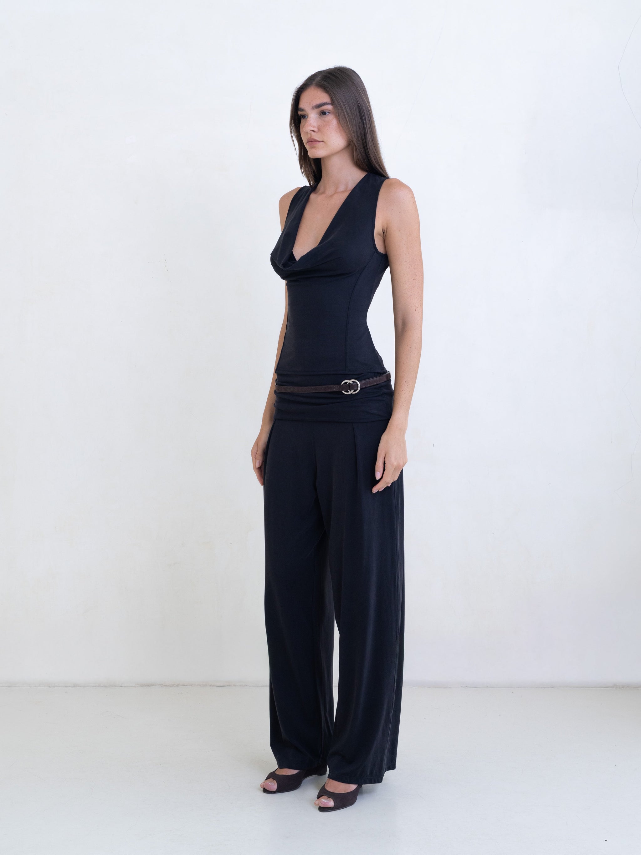 Woman wearing a black sleeveless cowl neck top and black pants on a white background. 