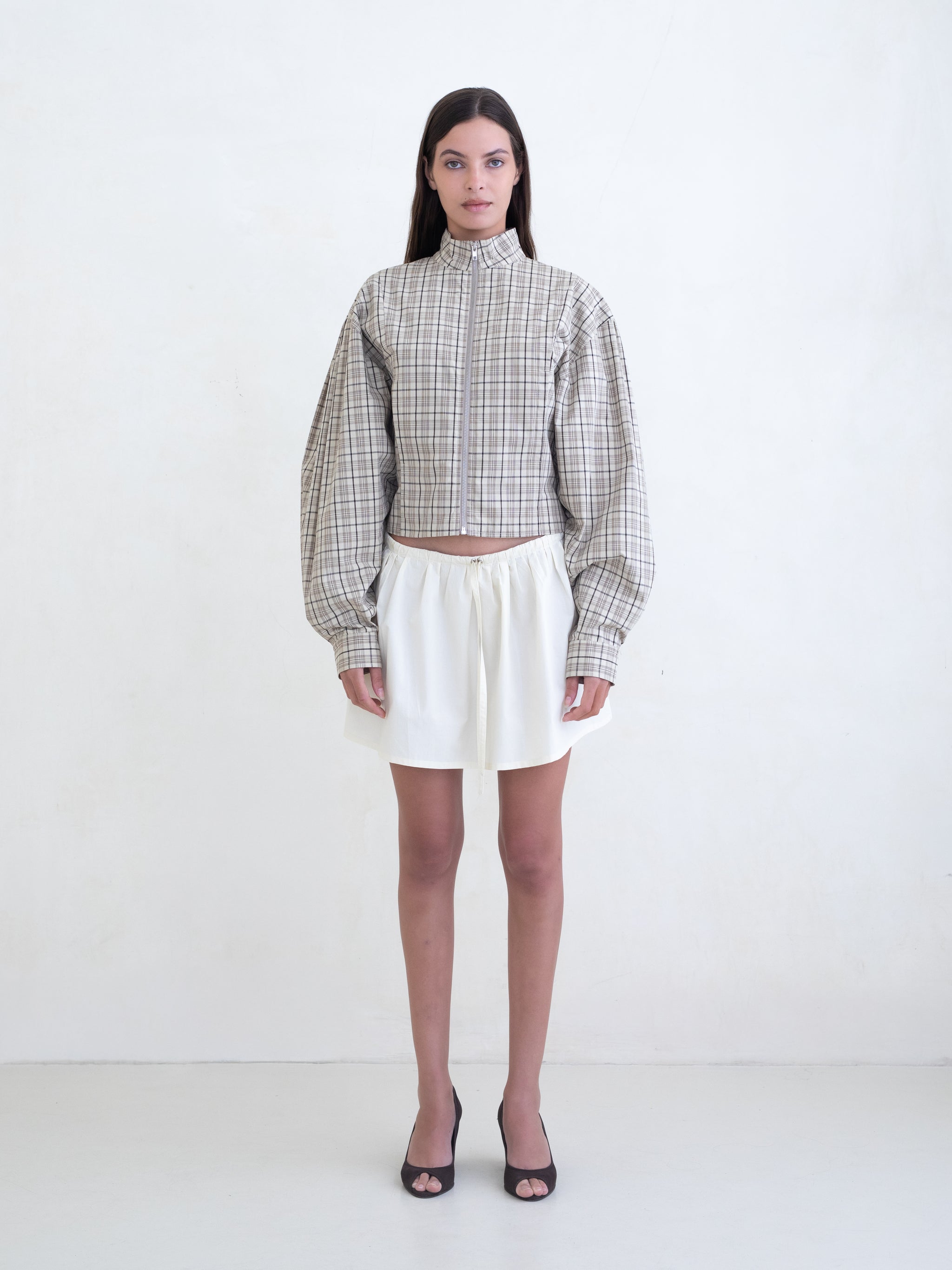 Woman wearing a brown plaid zip-front jacket and a cream mini skirt on a white background. 