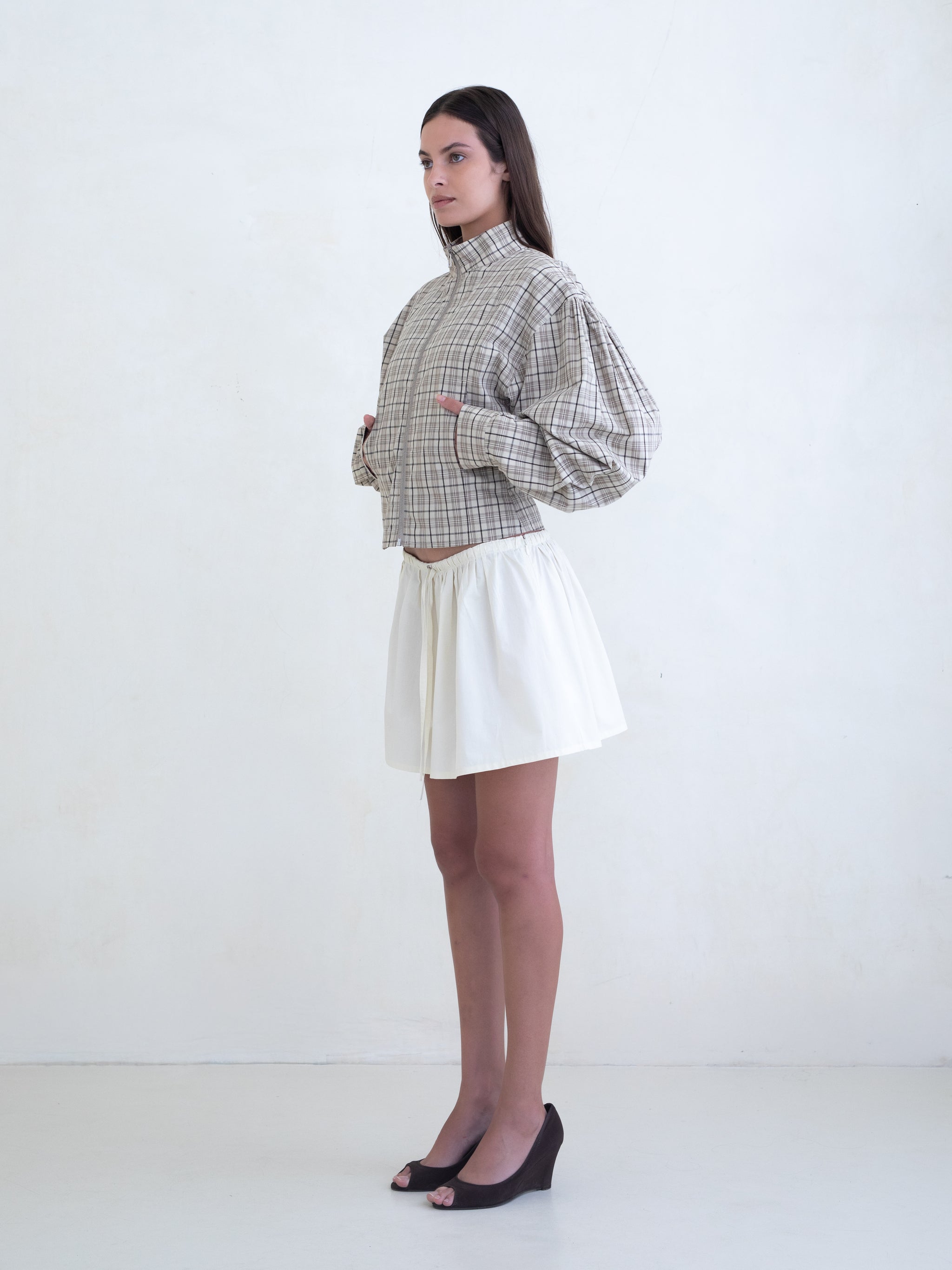 Woman wearing a brown plaid zip-front jacket and a cream mini skirt on a white background. 