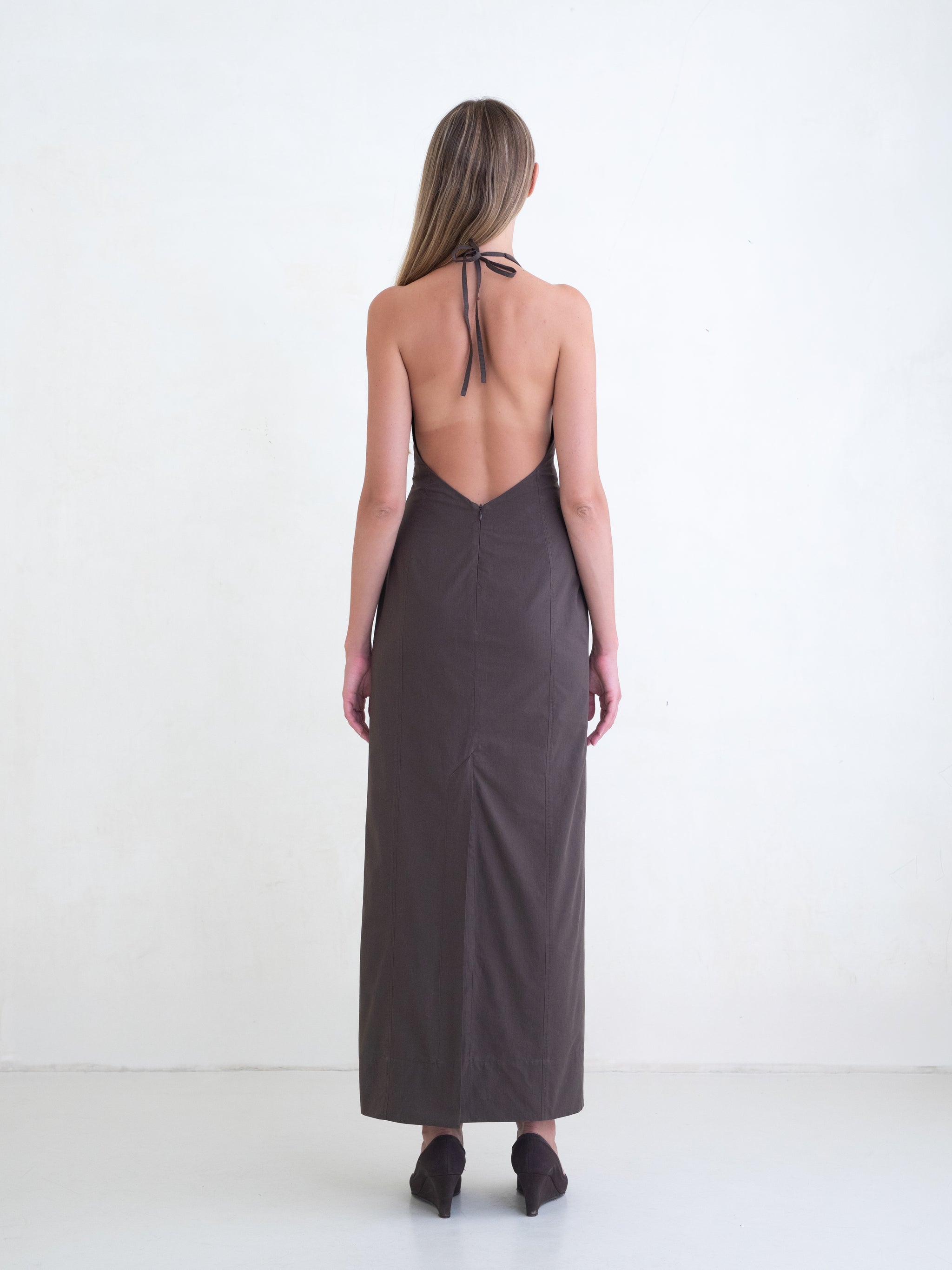 Woman wearing a brown sleeveless maxi dress with open low back on a white background. 