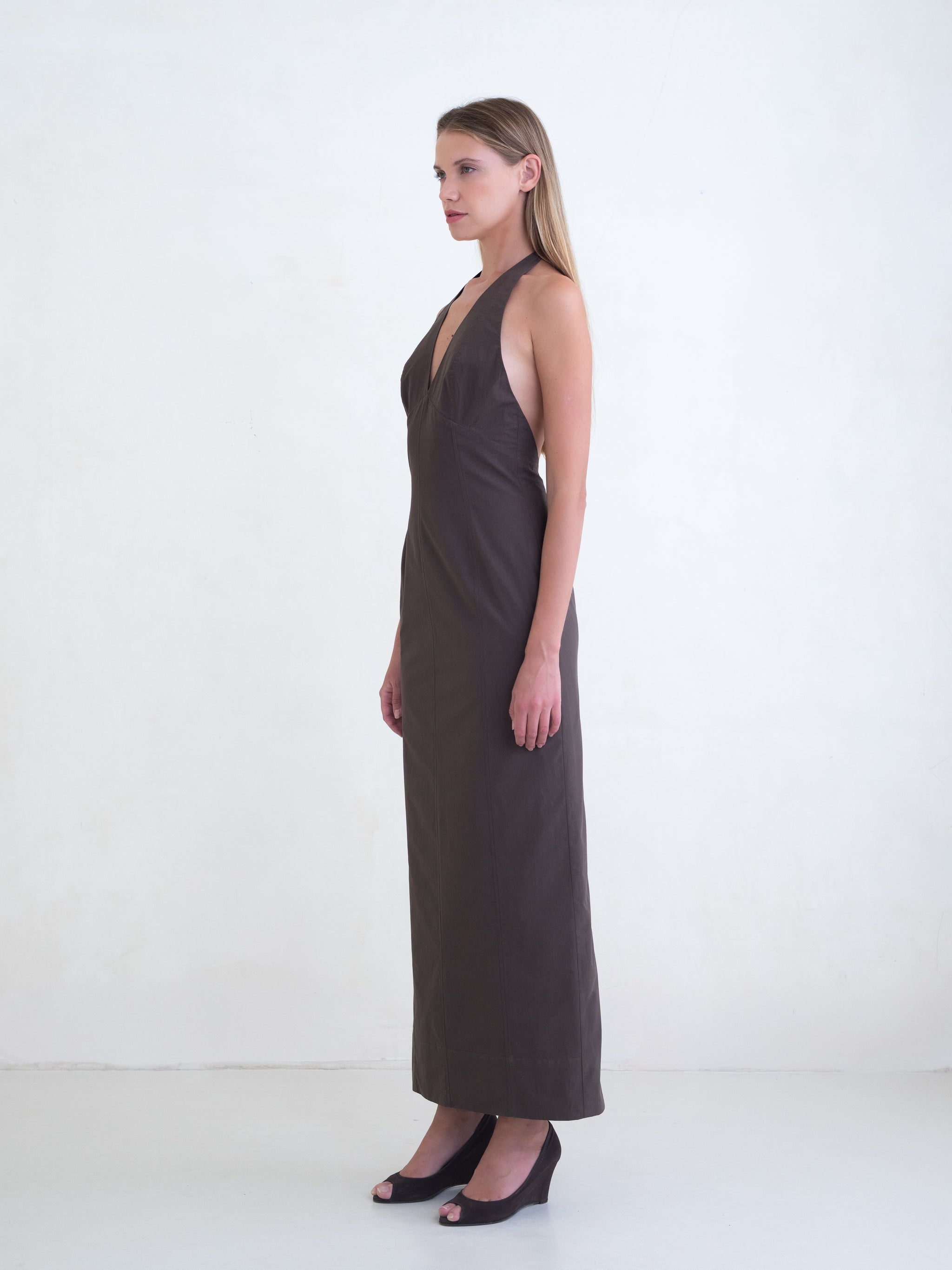 Woman wearing a brown sleeveless maxi dress with open low back on a white background. 