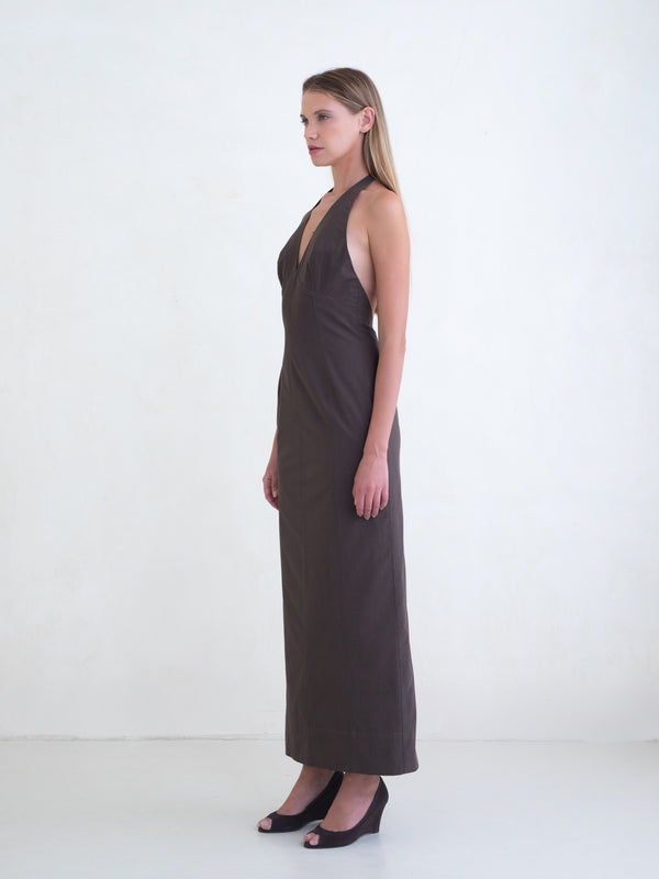 Woman wearing a brown sleeveless maxi dress with open low back on a white background. 