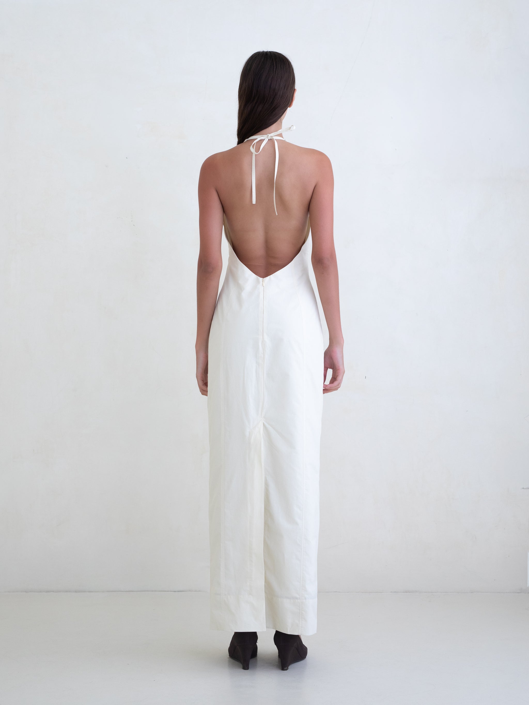 Woman wearing a cream sleeveless maxi dress with open low back on a white background. 