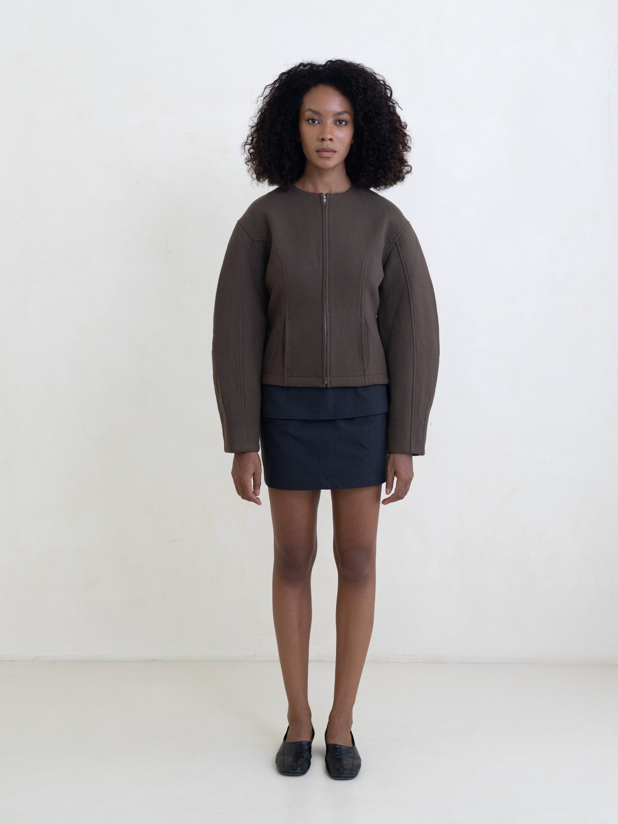 Woman wearing a brown zip-front jacket and a black mini skirt on a white background. 