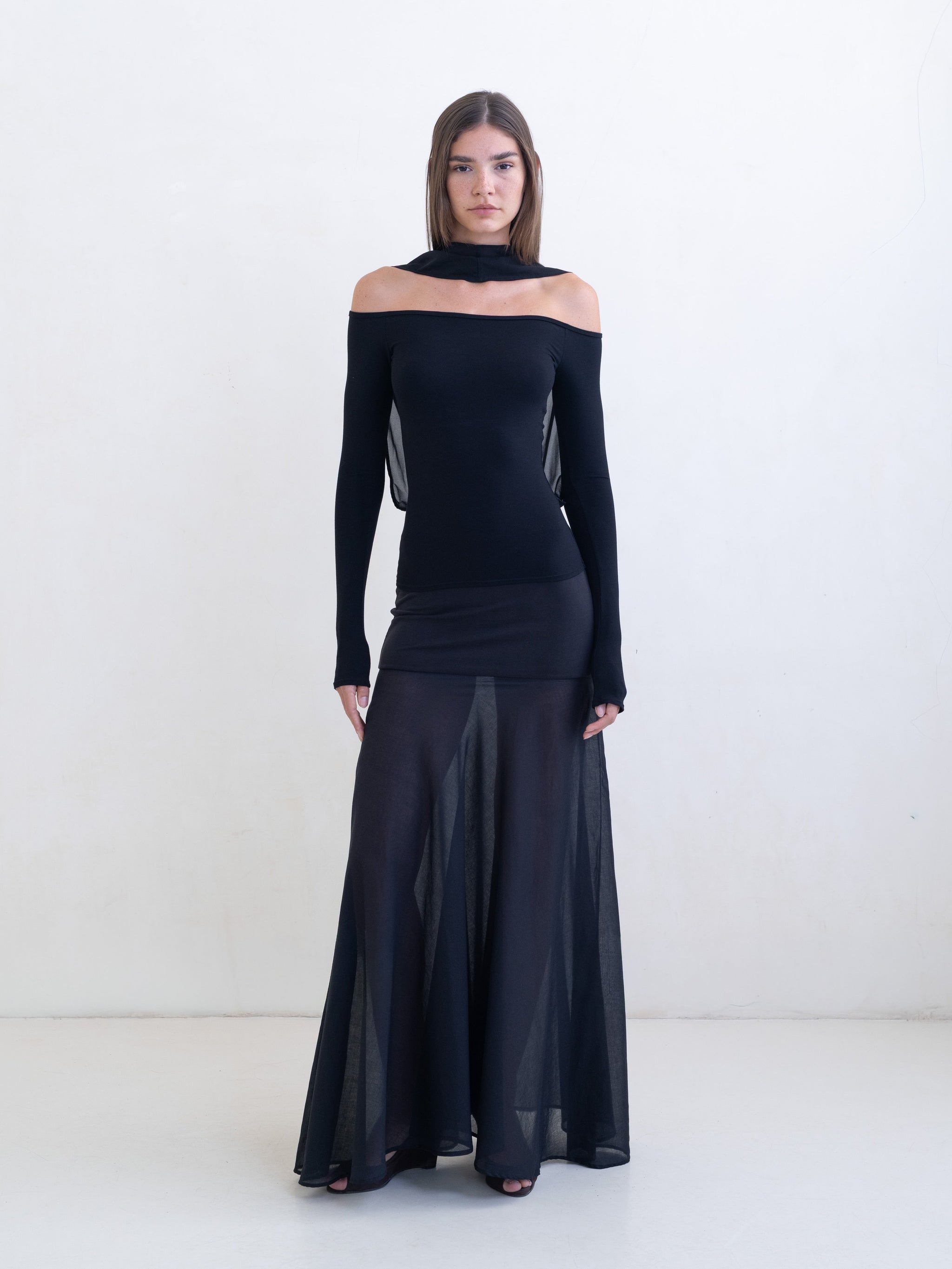 Woman wearing a black off-the-shoulder top and a sheer black maxi skirt on a white background.