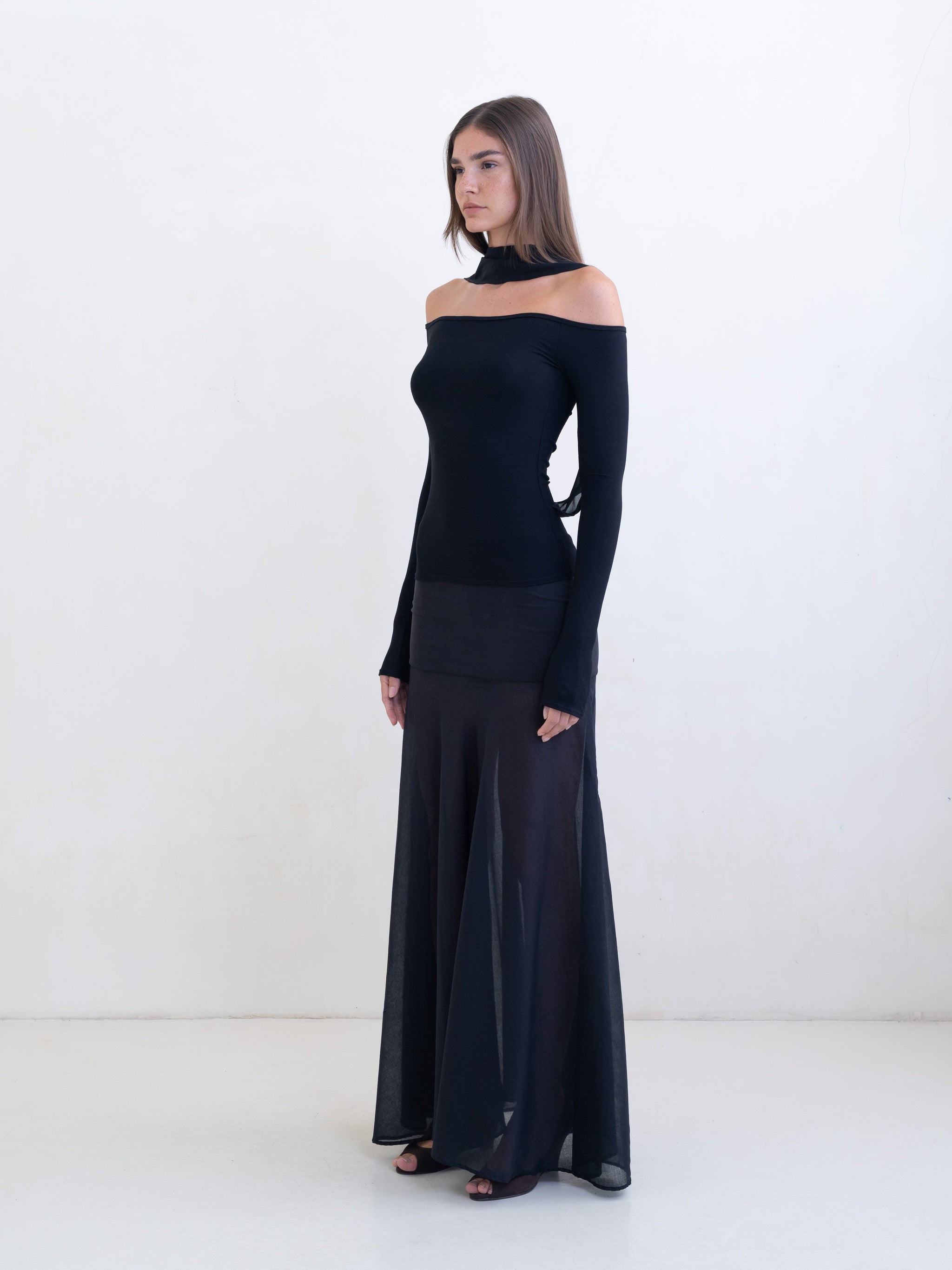 Woman wearing a black off-the-shoulder top and a sheer black maxi skirt on a white background.