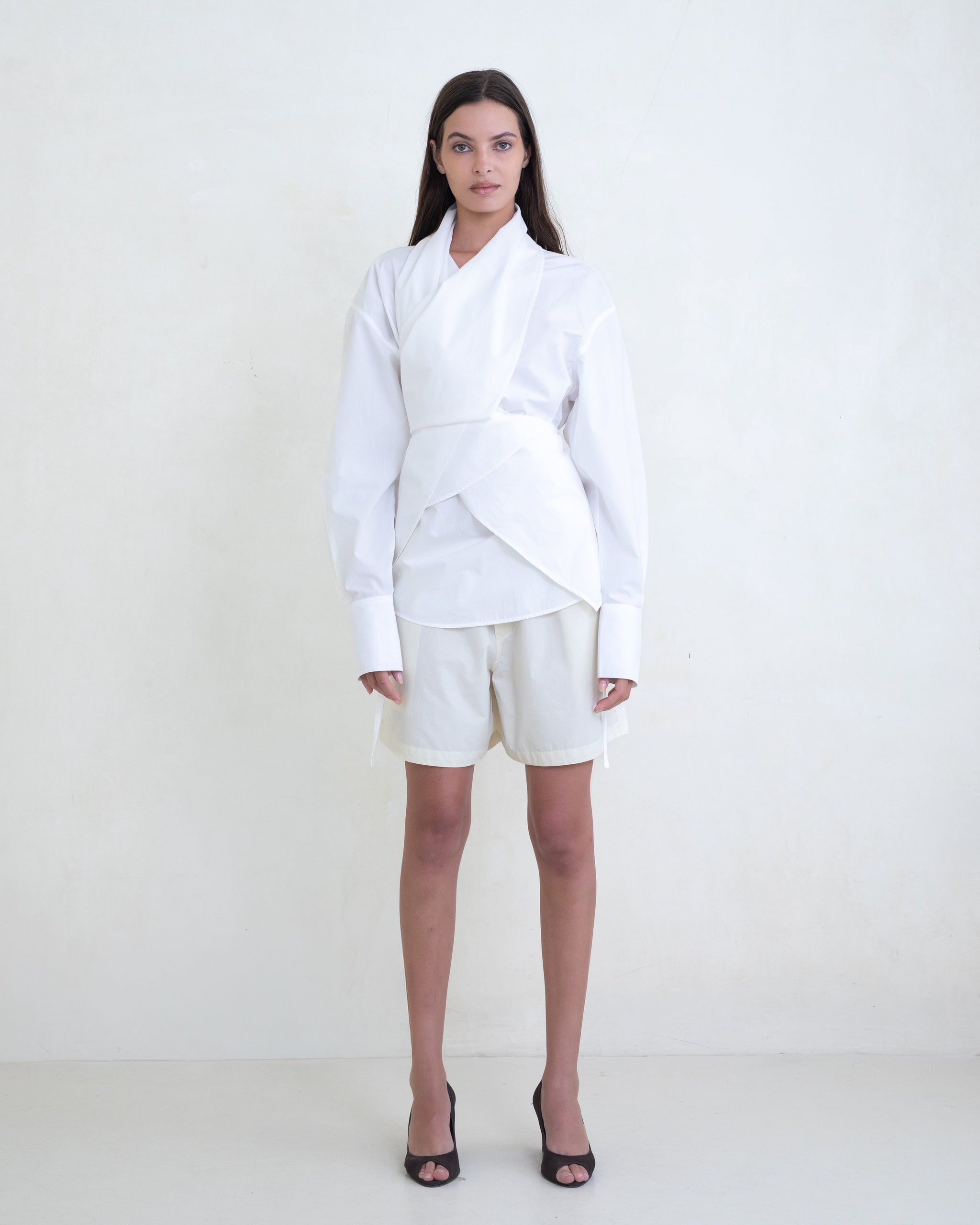 Woman wearing a white wrap shirt and cream shorts on a white background. 