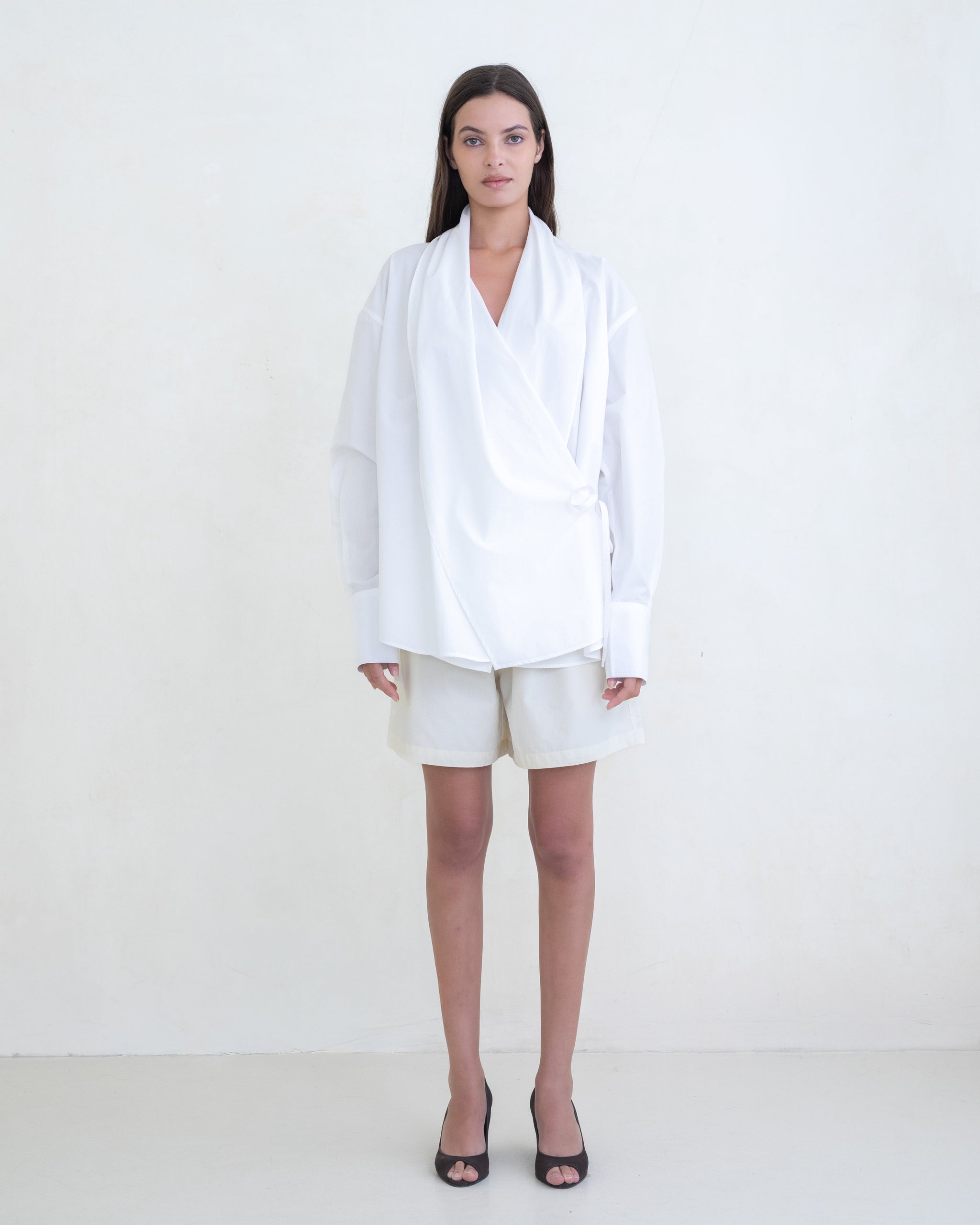 Woman wearing a white wrap shirt and cream shorts on a white background.  