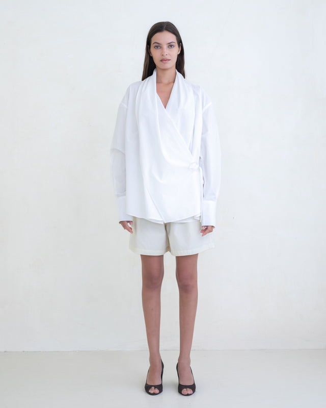 Woman wearing a white wrap shirt and cream shorts on a white background.  