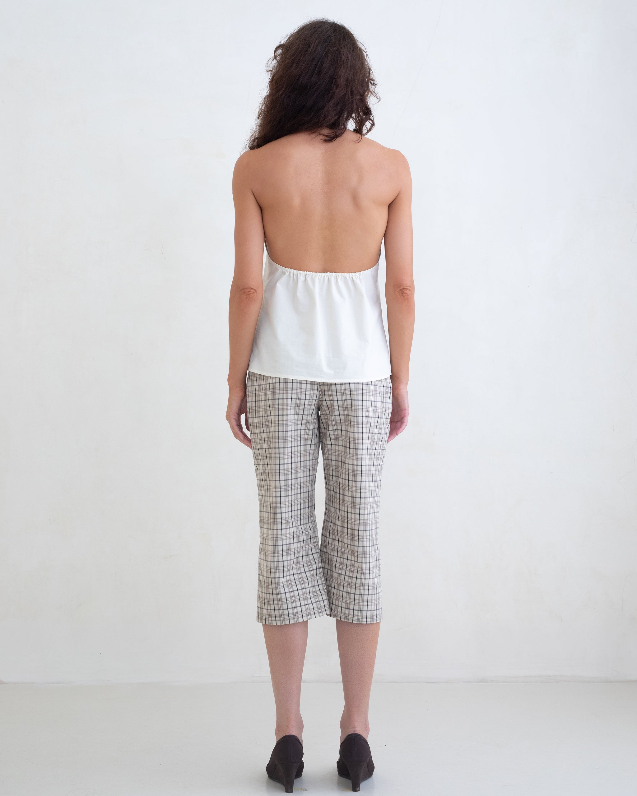 Woman wearing a cream v-neck halter top and brown plaid capri pants on a white background. 