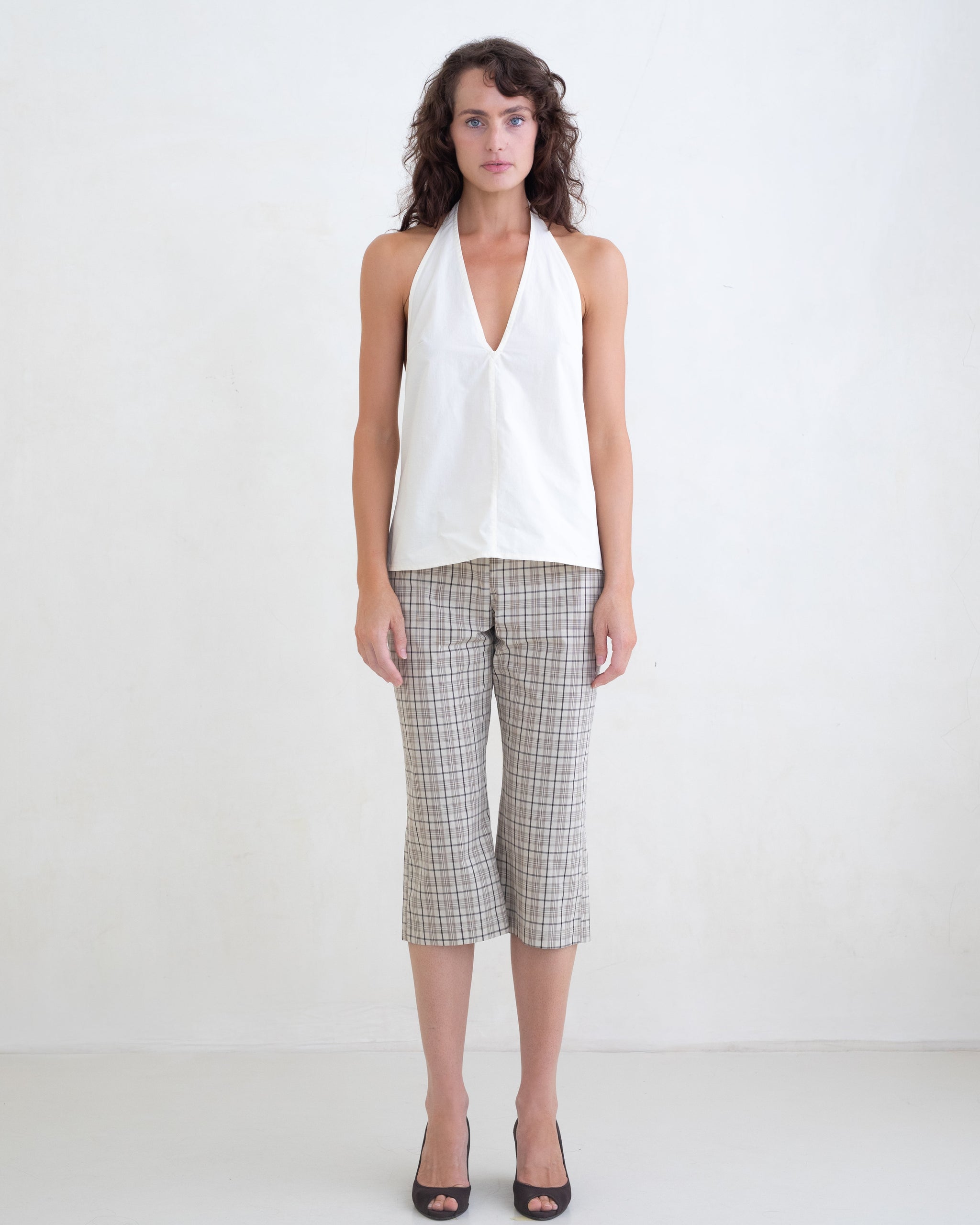 Woman wearing a cream v-neck halter top and brown plaid capri pants on a white background. 