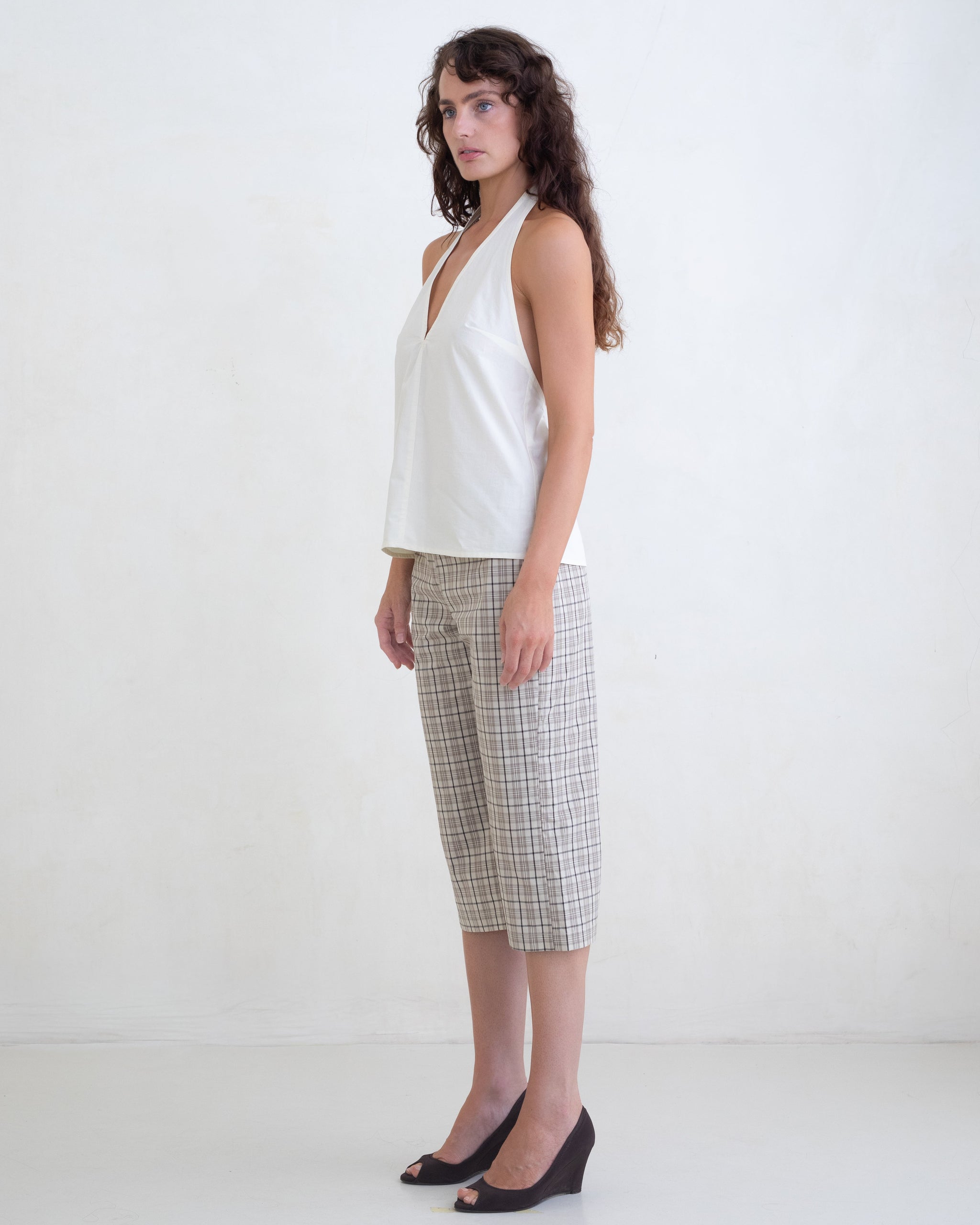 Woman wearing a cream v-neck halter top and brown plaid capri pants on a white background. 