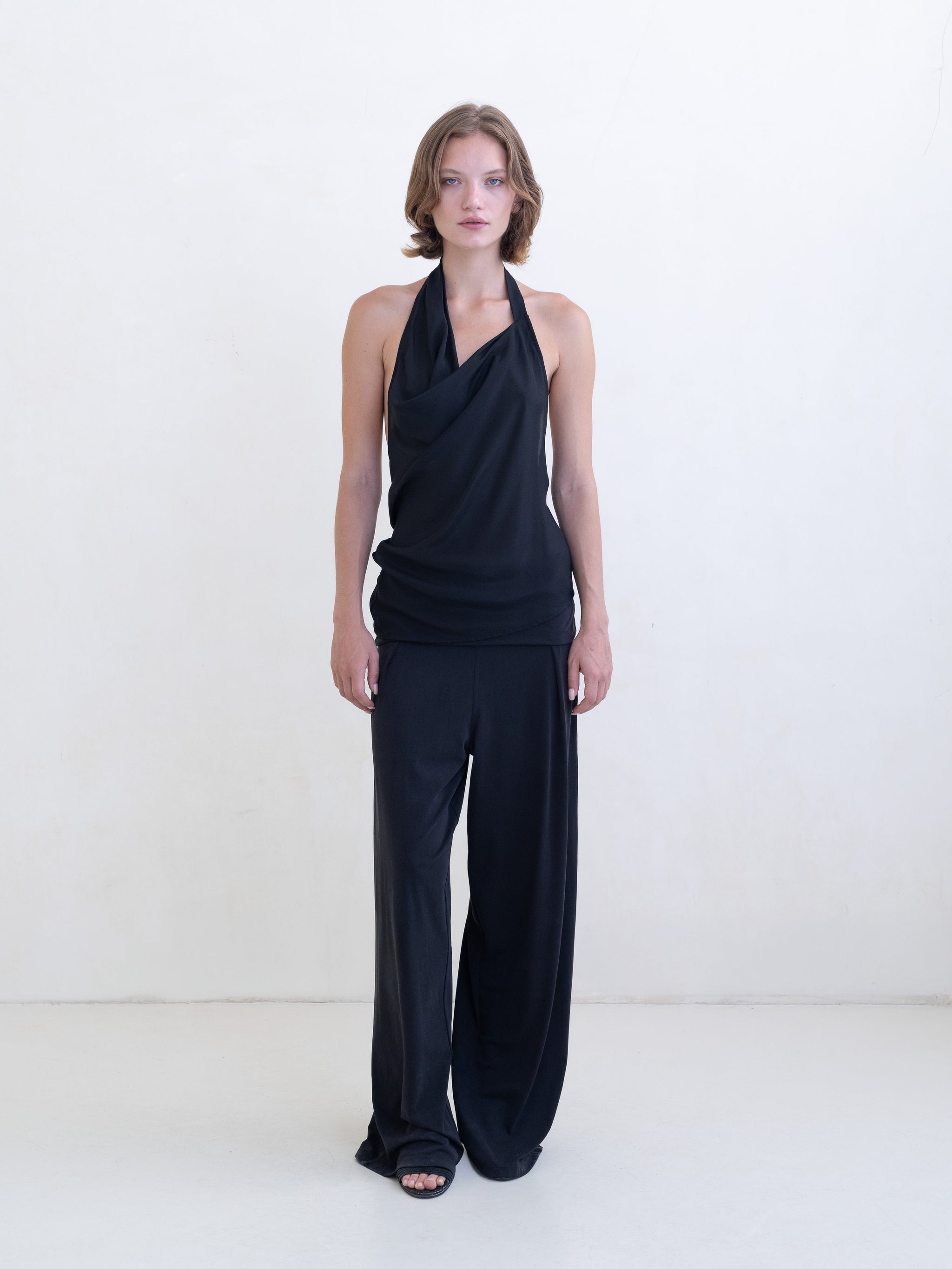 Woman wearing a black asymmetric halter-neck top and black pants on a white background. 
