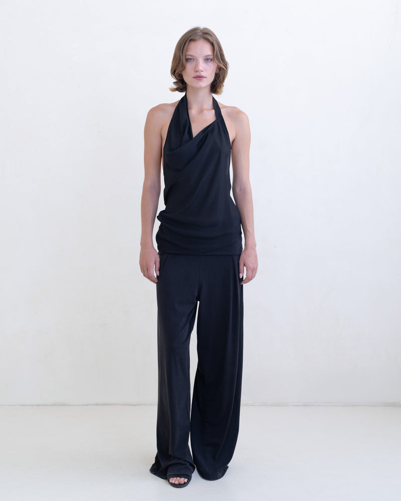 Woman wearing a black asymmetric halter-neck top and black pants on a white background. 