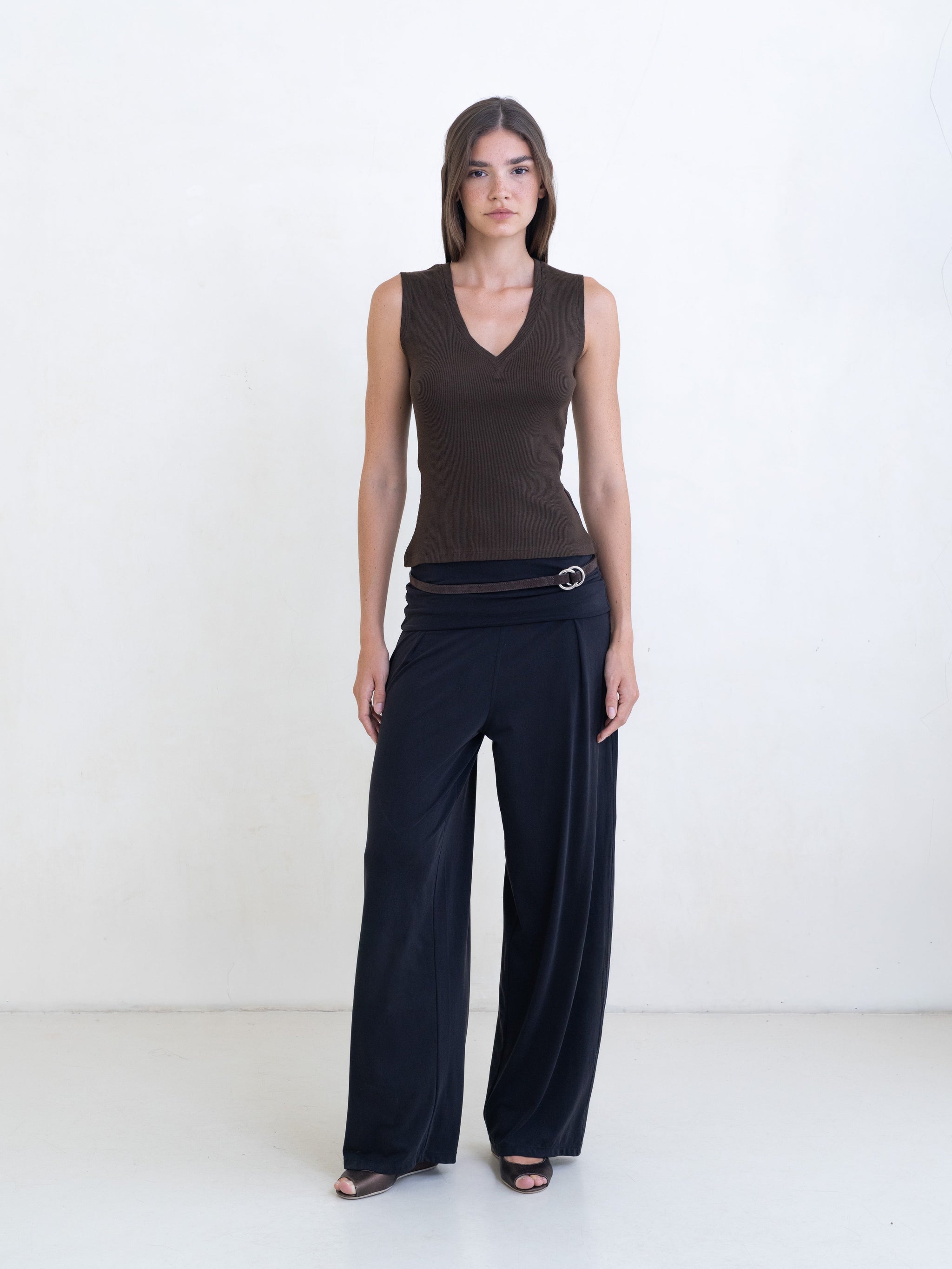 Woman wearing a brown sleeveless v-neck top and black pants on a white background. 
