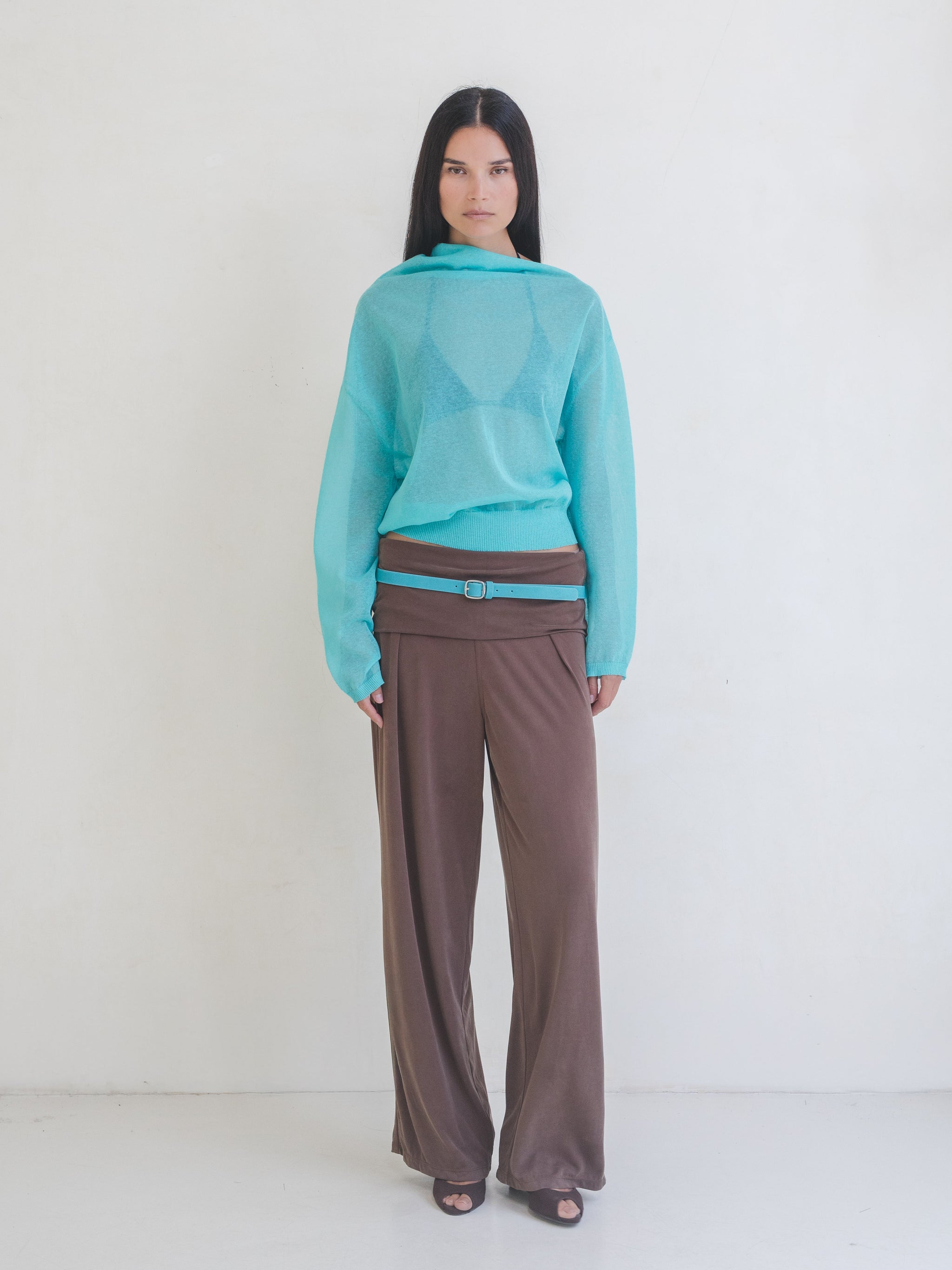 Woman wearing a bright blue sweater, brown pants with a bright blue leather belt on a white background. 