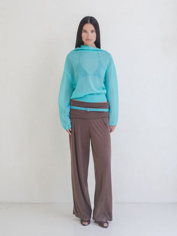 Woman wearing a bright blue sweater, brown pants with a bright blue leather belt on a white background. 