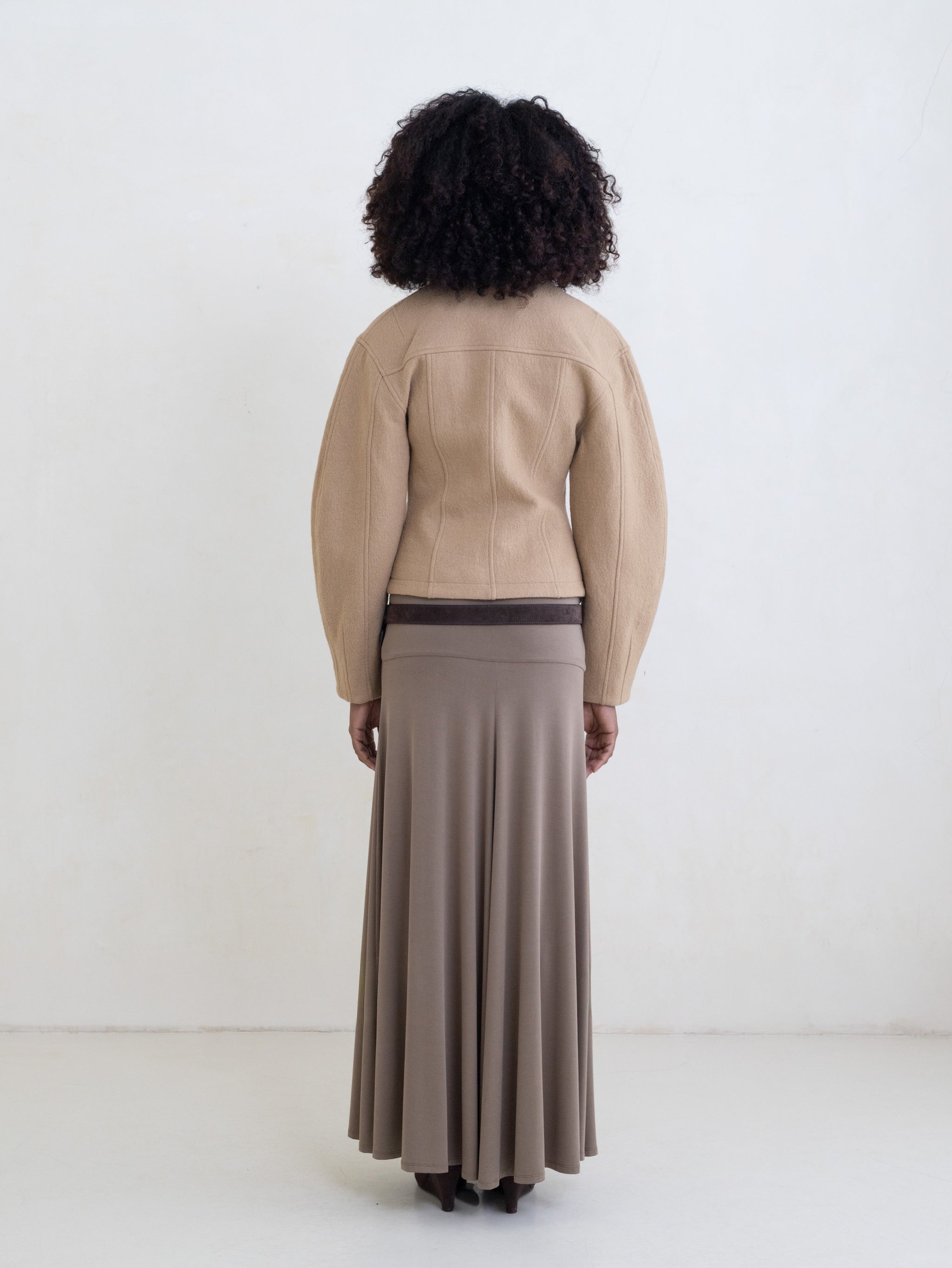 Woman wearing a beige jacket and a taupe maxi skirt with a brown leather belt on a white background.
