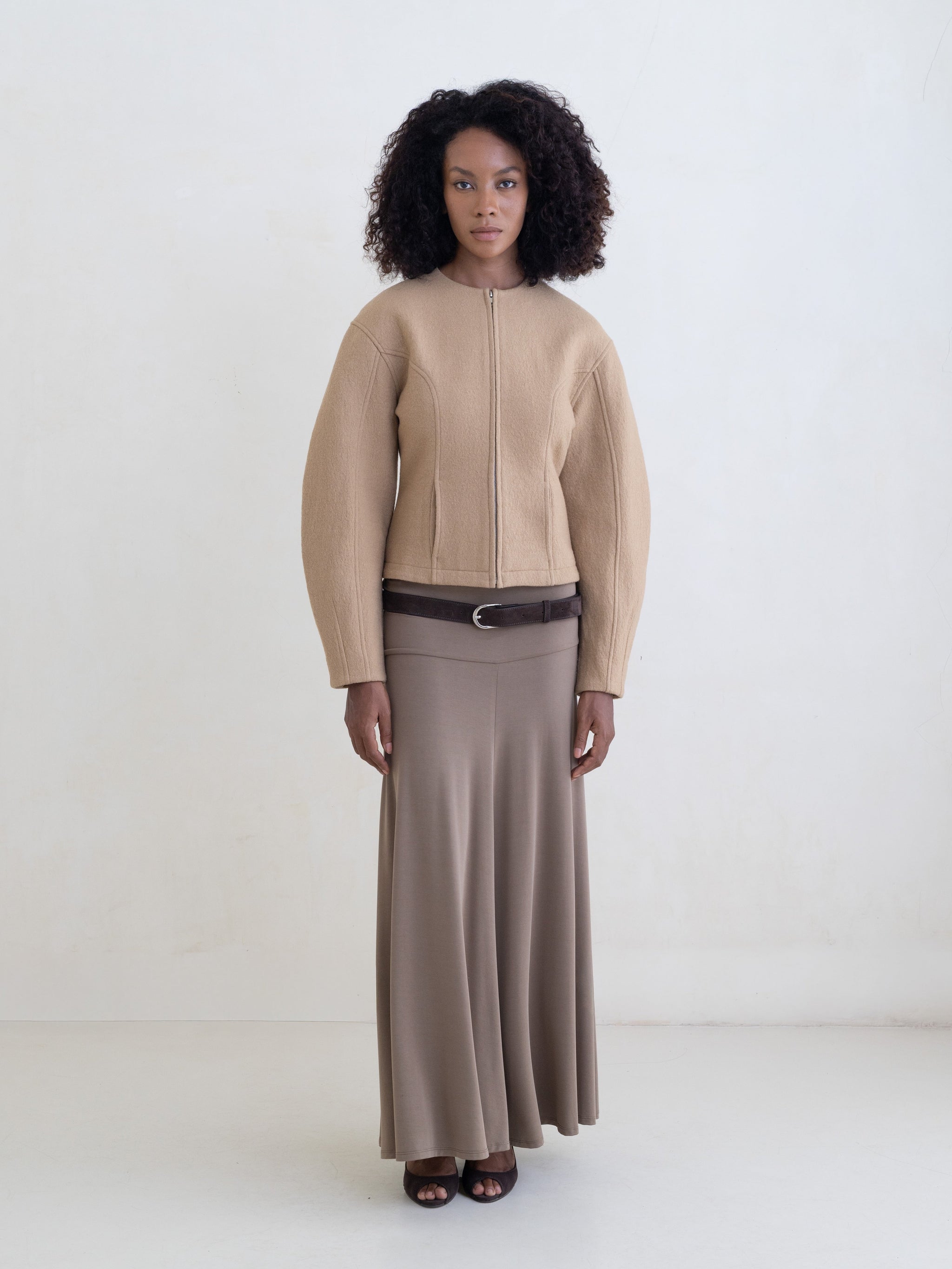 Woman wearing a beige jacket and a taupe maxi skirt with a brown leather belt on a white background.