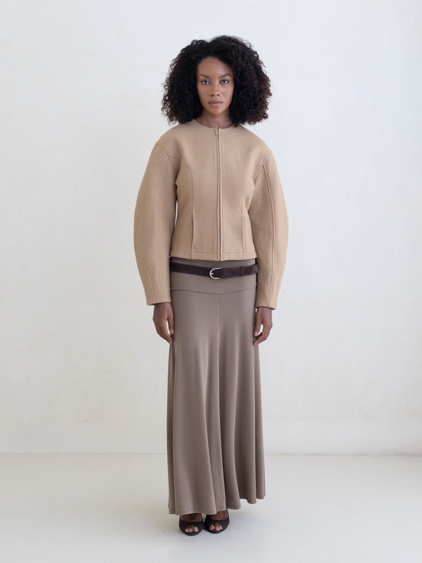 Woman wearing a beige jacket and a taupe maxi skirt with a brown leather belt on a white background.