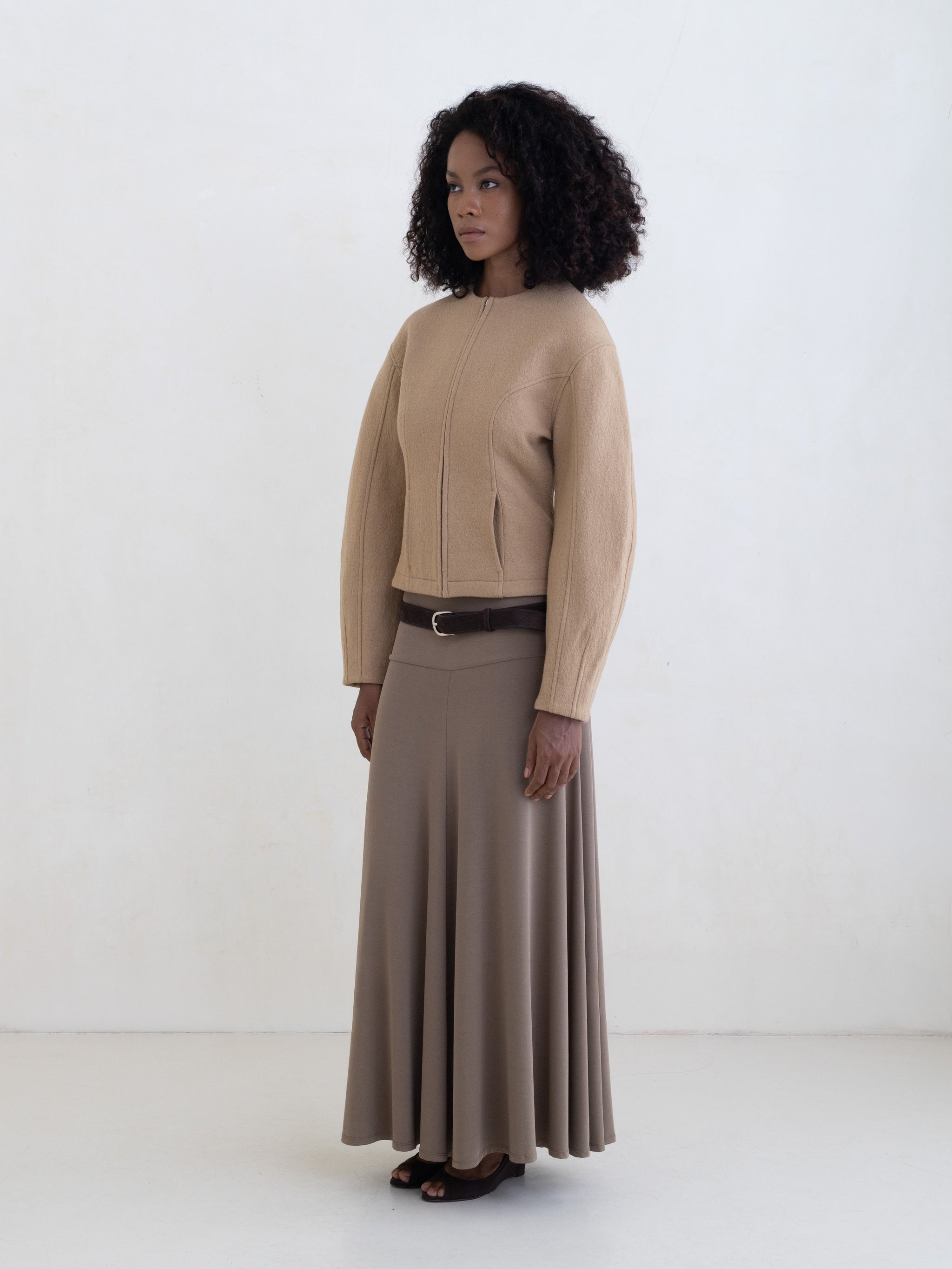 Woman wearing a beige jacket and a taupe maxi skirt with a brown leather belt on a white background.